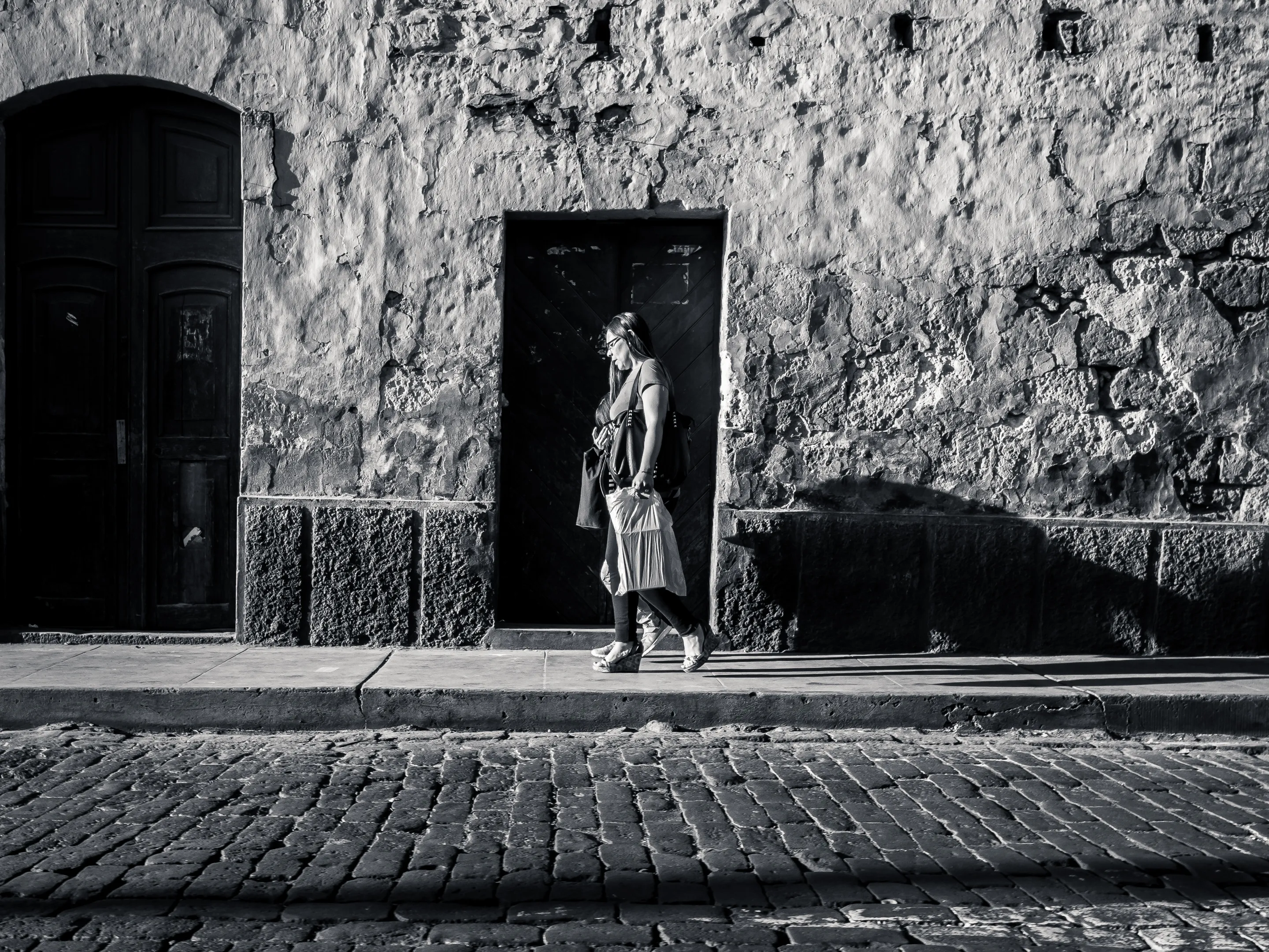 Person exiting stone building doorway, carrying a bag, standing on cobblestone street, black and white.