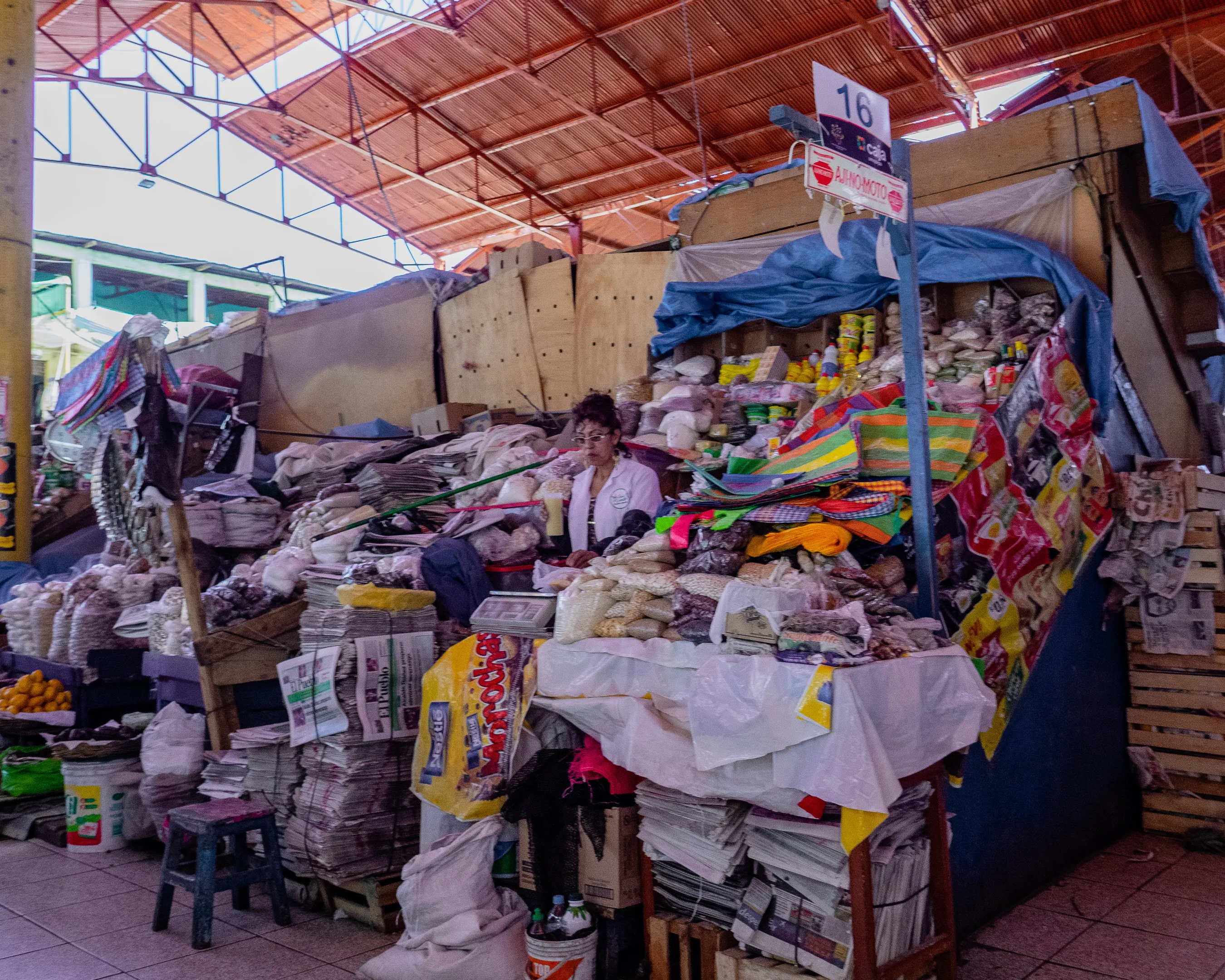A bustling market stall with a variety of colorful textiles and bags, a man sitting on a stool surrounded by goods, under a corrugated metal roof.