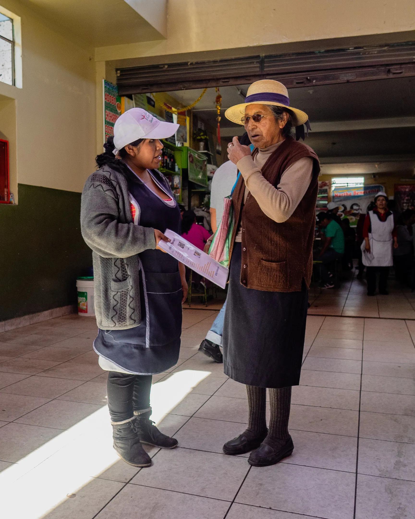 Two women standing indoors, one holding a microphone, the other holding papers, both wearing hats and traditional clothing.