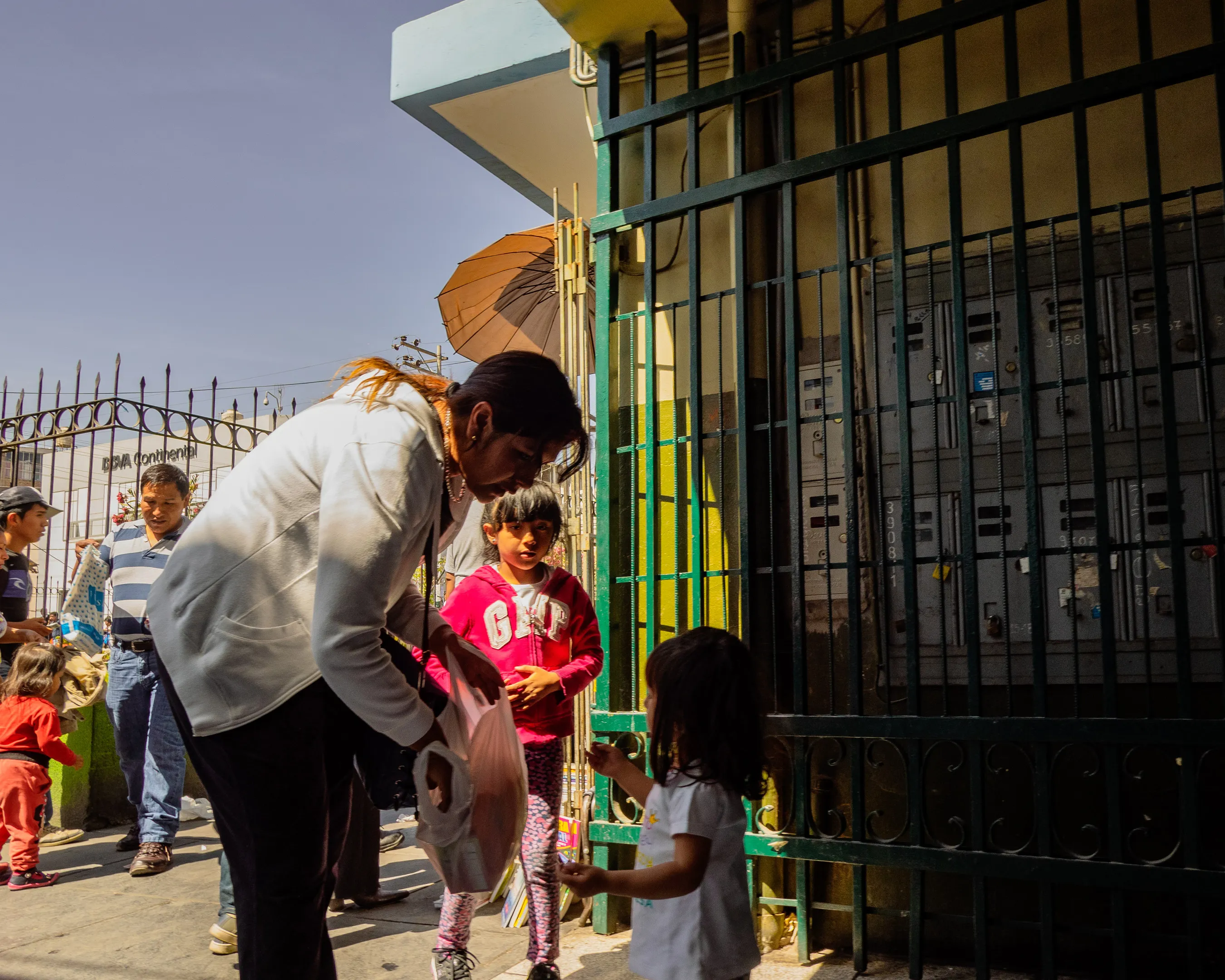 Woman assisting child at a gated entrance, others waiting in background under clear sky.
