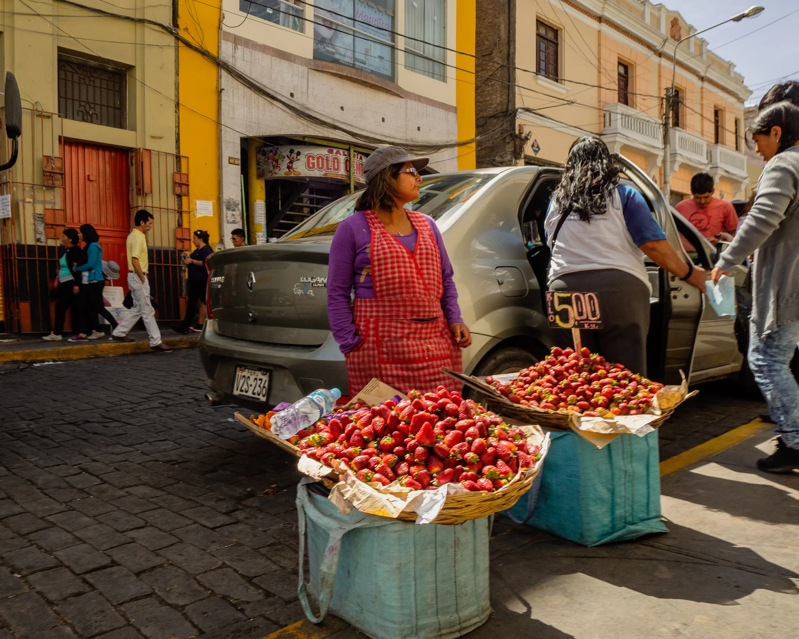 Street vendor selling red apples from baskets in front of a yellow building, with a parked car and customers nearby.