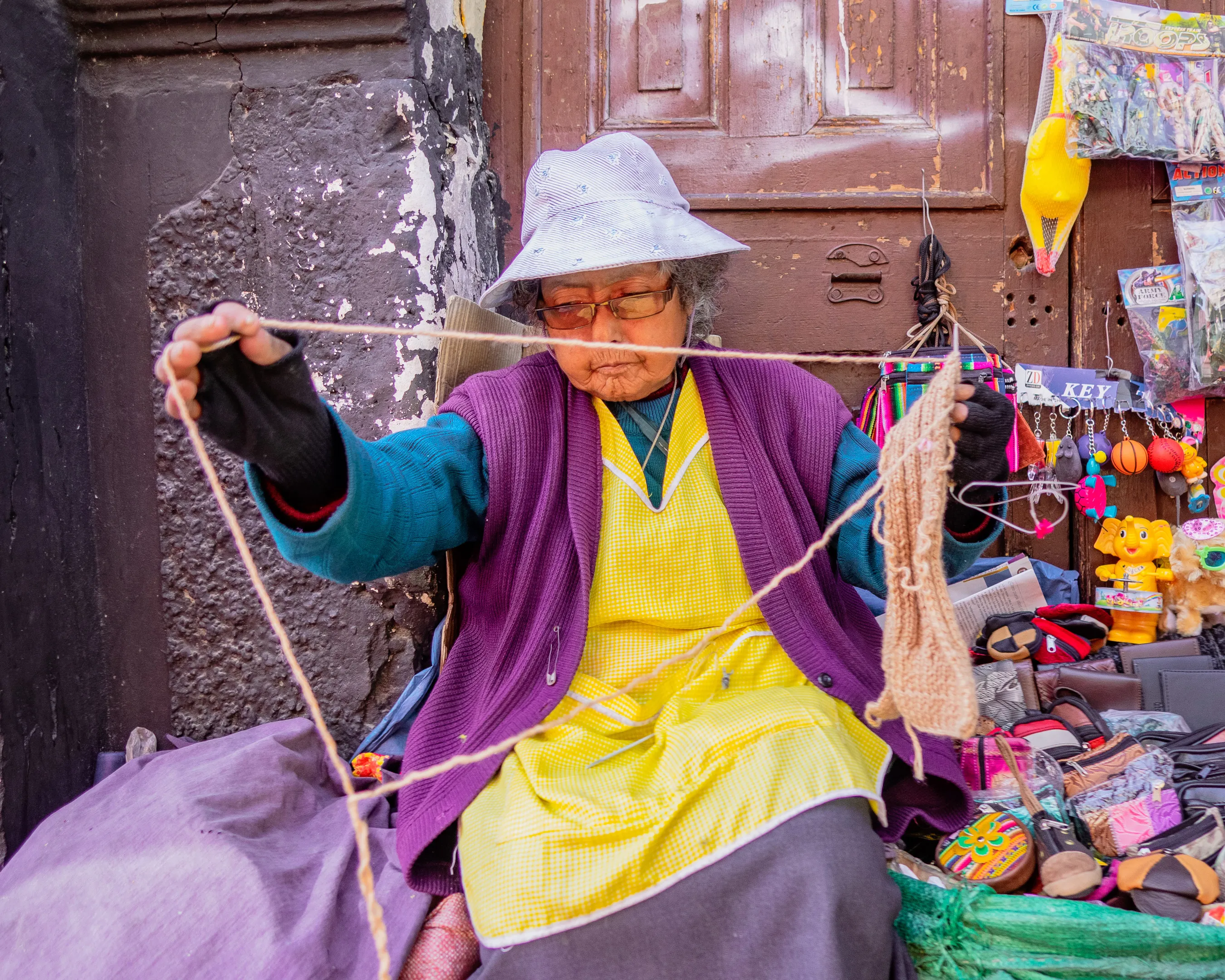 Elderly woman in white hat weaving on a street side stall with colorful items.