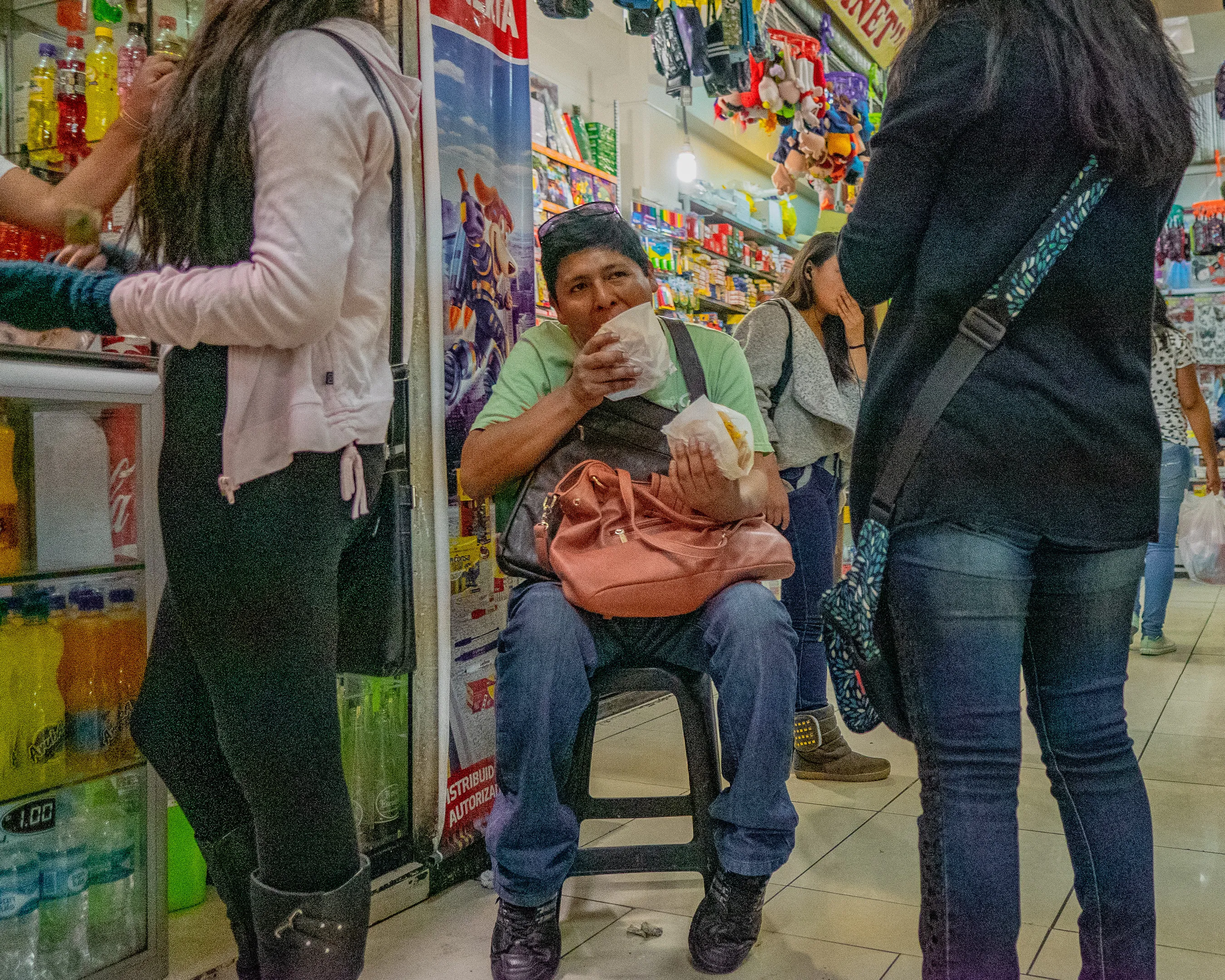 A man sitting on a stool in a store, eating from a bag and holding a drink.