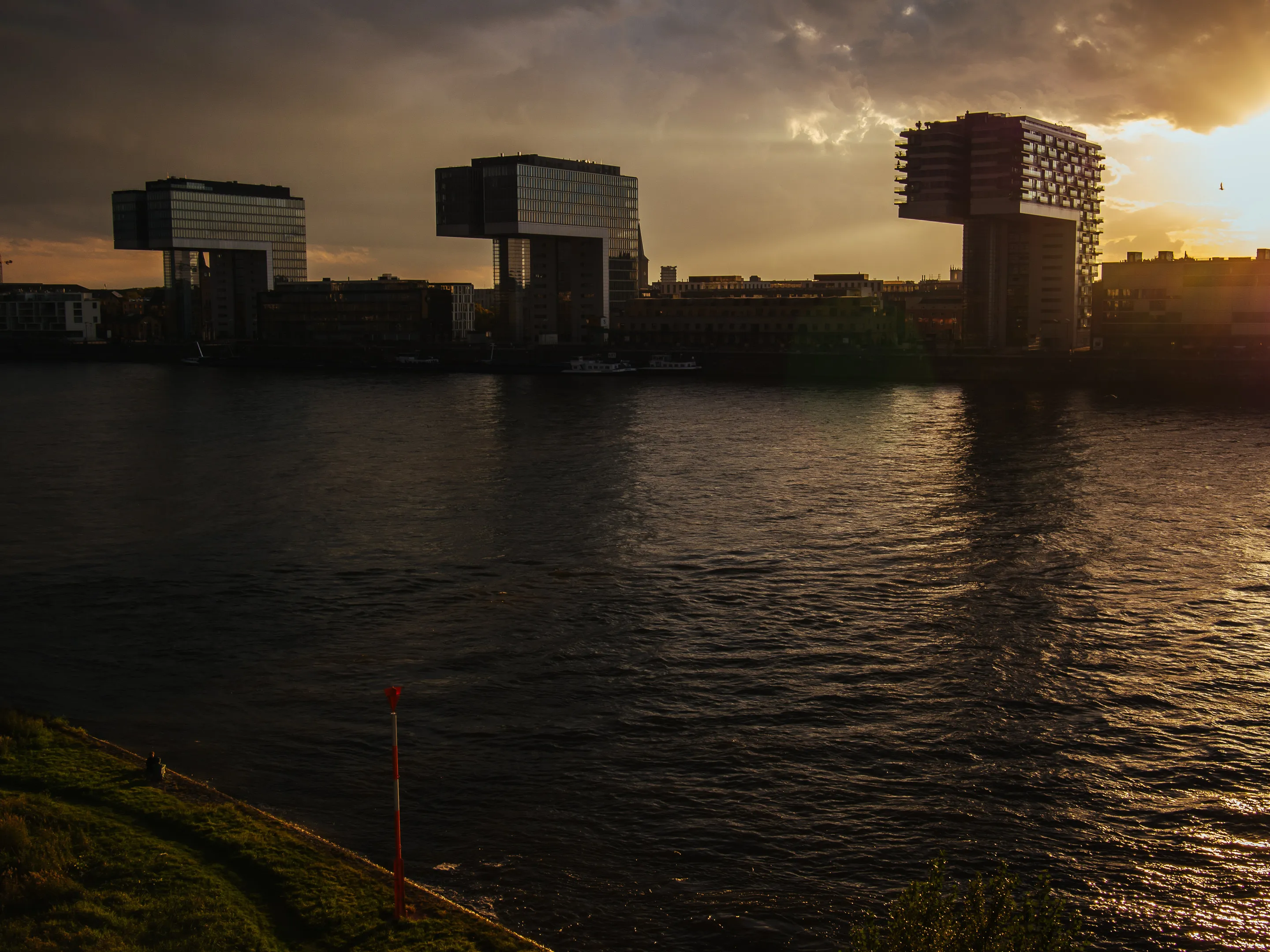 Three modern buildings with unique, cube-shaped designs on a riverbank at sunset.