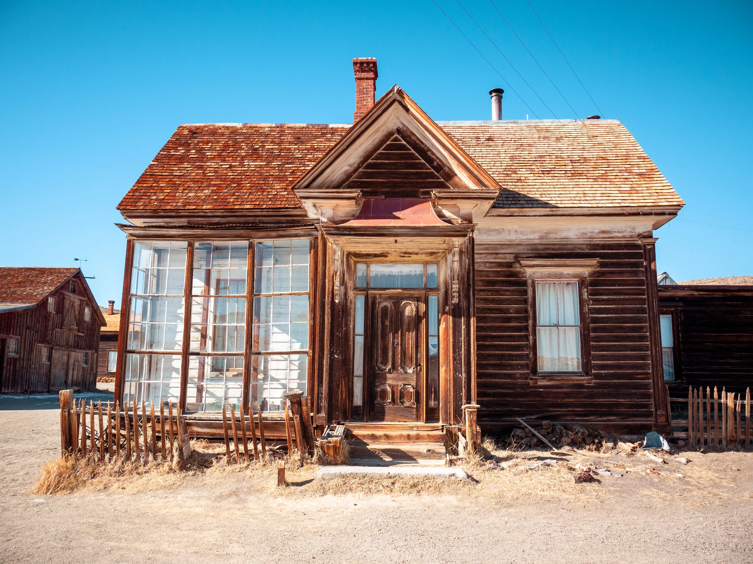 Abandoned wooden house with peeling paint, broken windows, and a dilapidated porch in a desert-like setting.