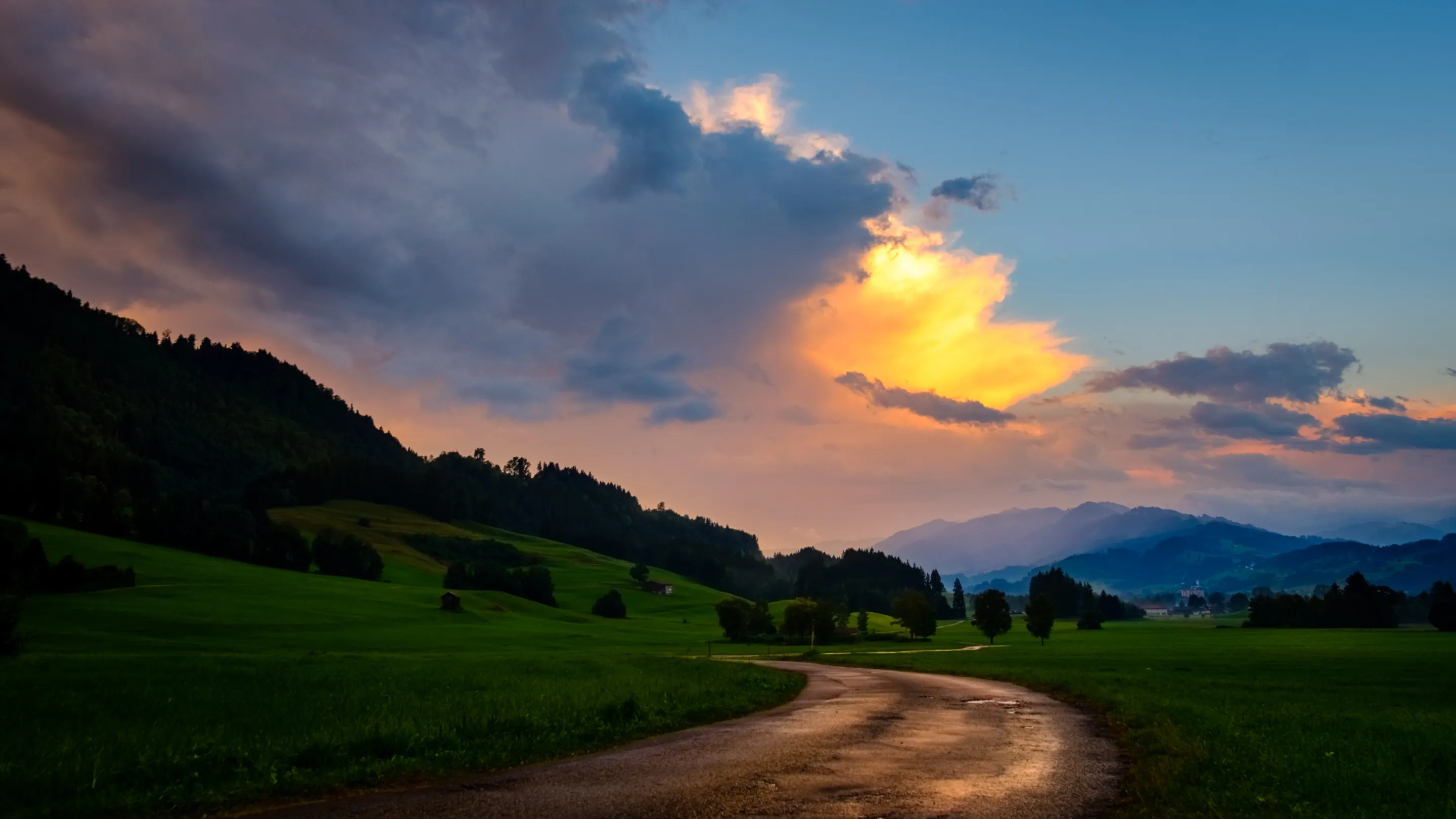 Sunset over a winding road in a green valley with mountains in the background, clouds illuminated by the setting sun.