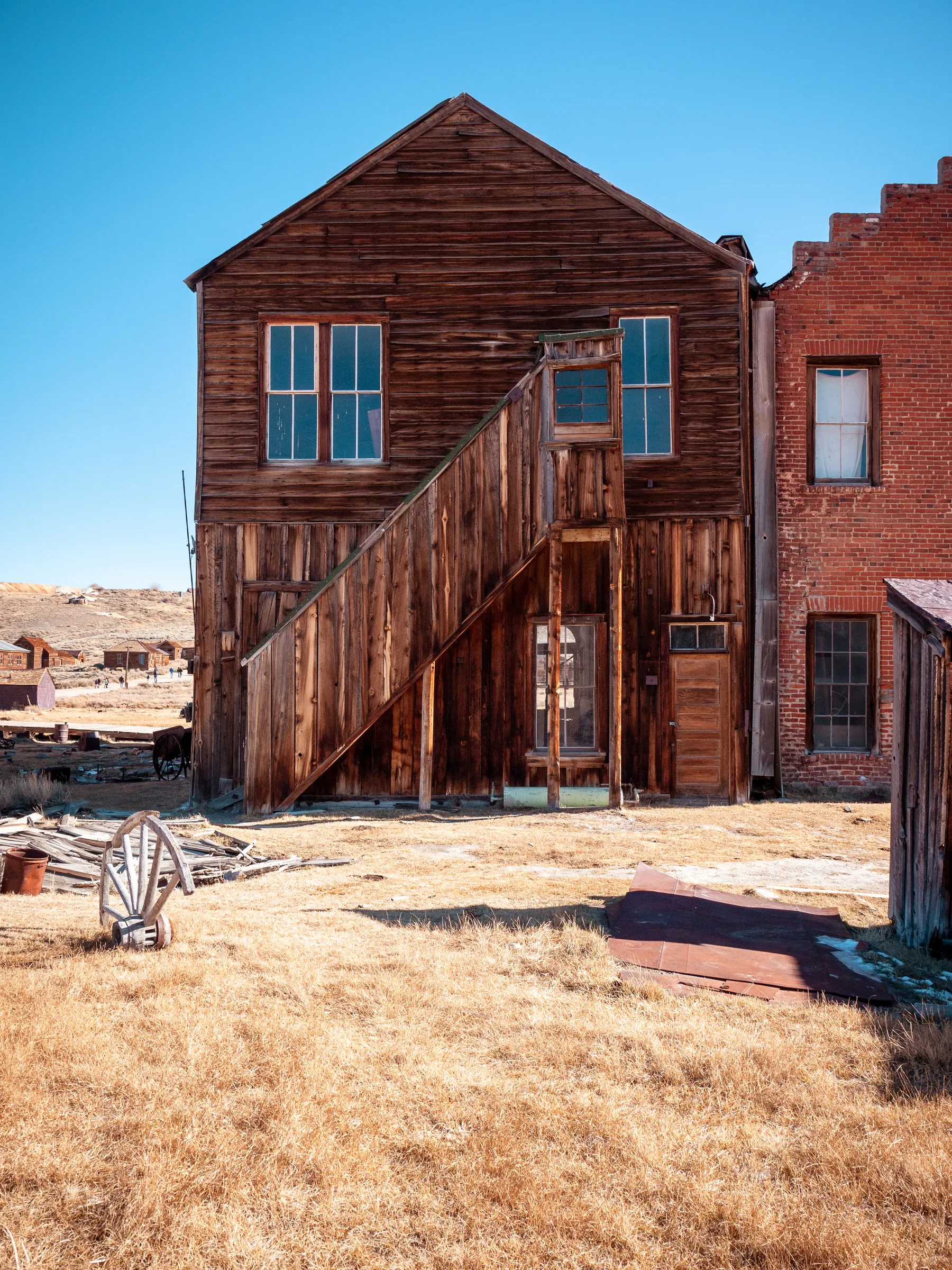 Weathered wooden building with a staircase leading to an entrance, surrounded by dry grass and other structures in a desert-like setting.