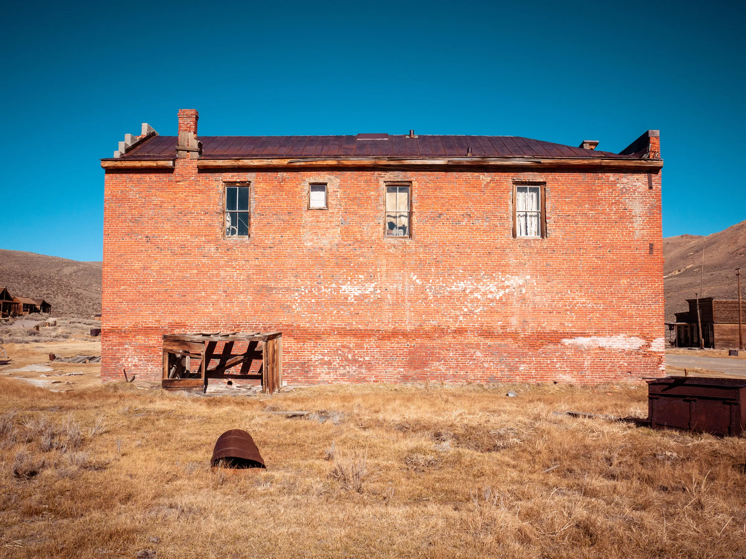 Abandoned red brick building with boarded-up windows in a dry, grassy landscape under clear blue sky.