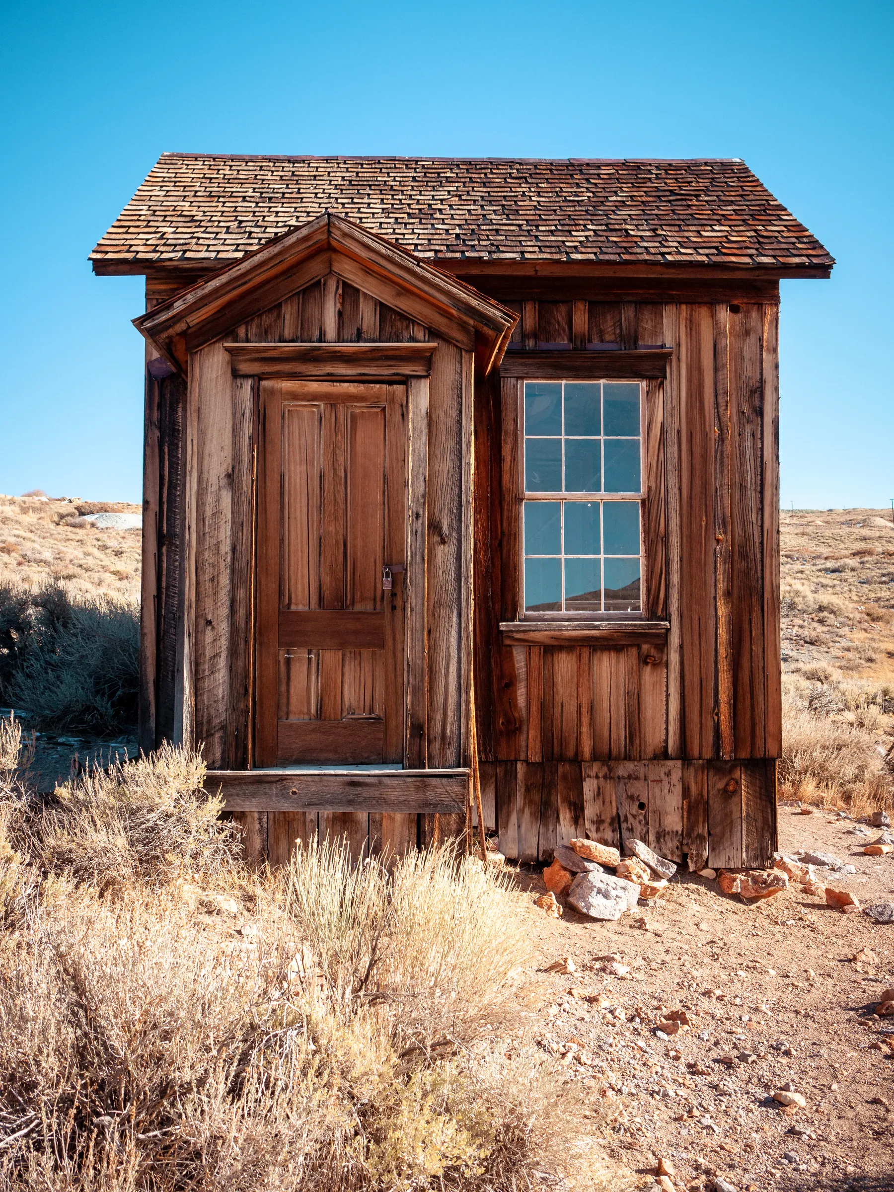 Weathered wooden cabin with a shingled roof, a door, and a window, set in a dry, grassy landscape.
