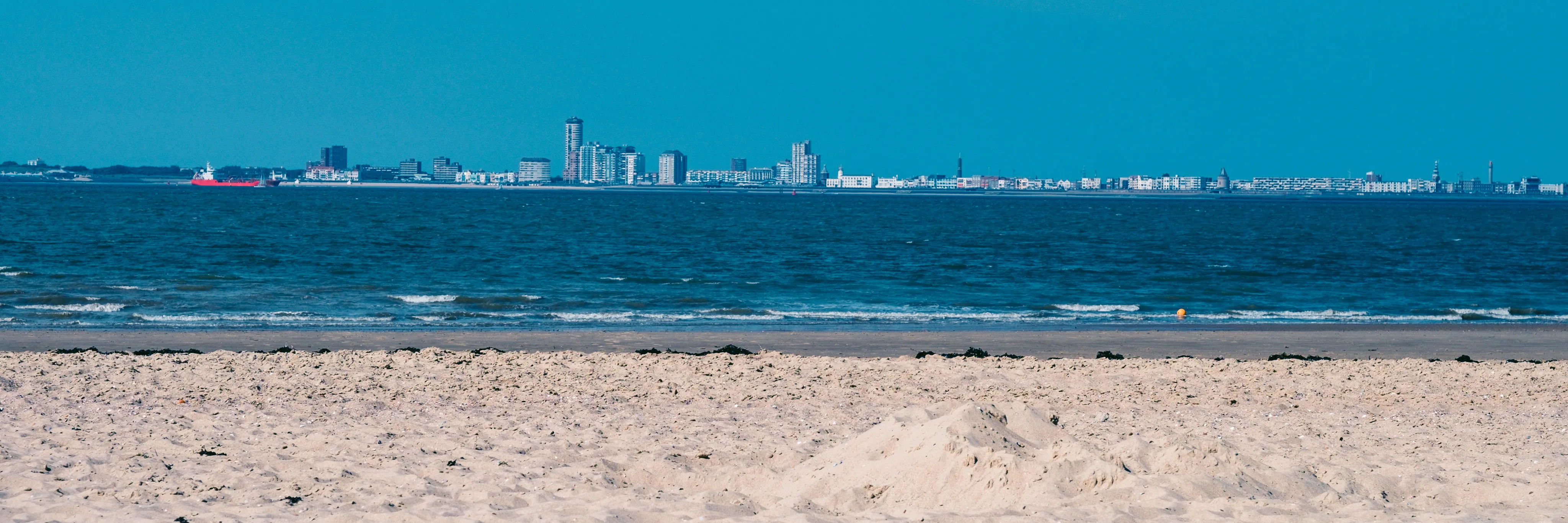 Panoramic view of an urban skyline across a calm blue sea, with a sandy beach in the foreground.