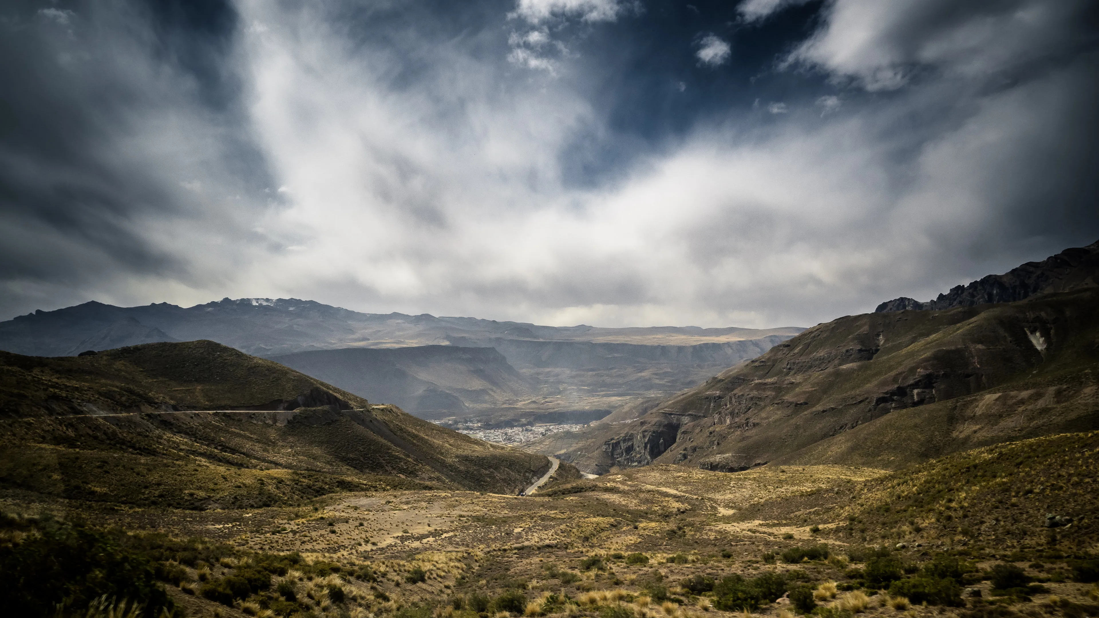 A winding road in a mountainous valley under dramatic, cloud-filled skies.