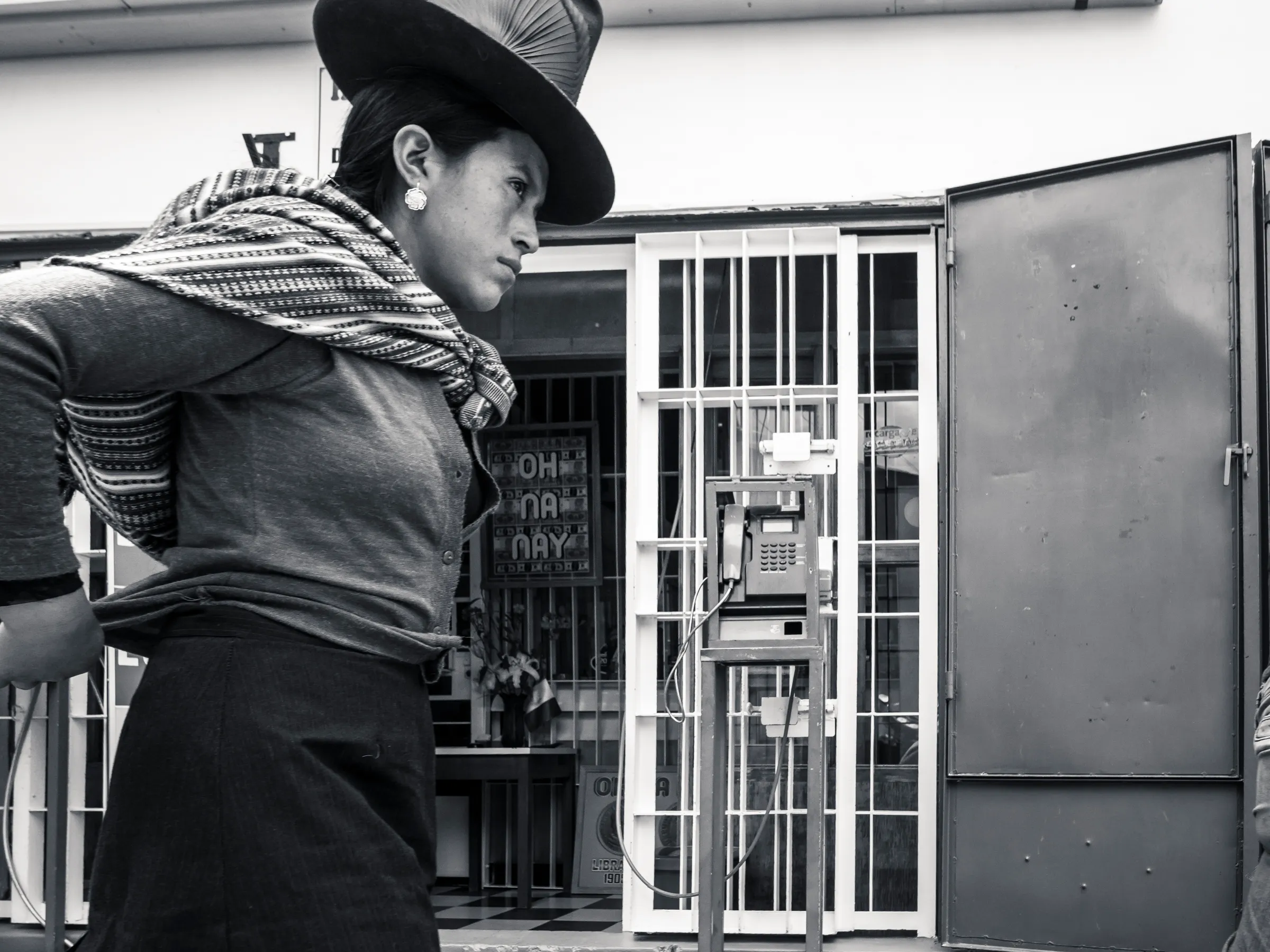 Person in traditional hat, scarf, looking away from a barred window with a telephone inside.