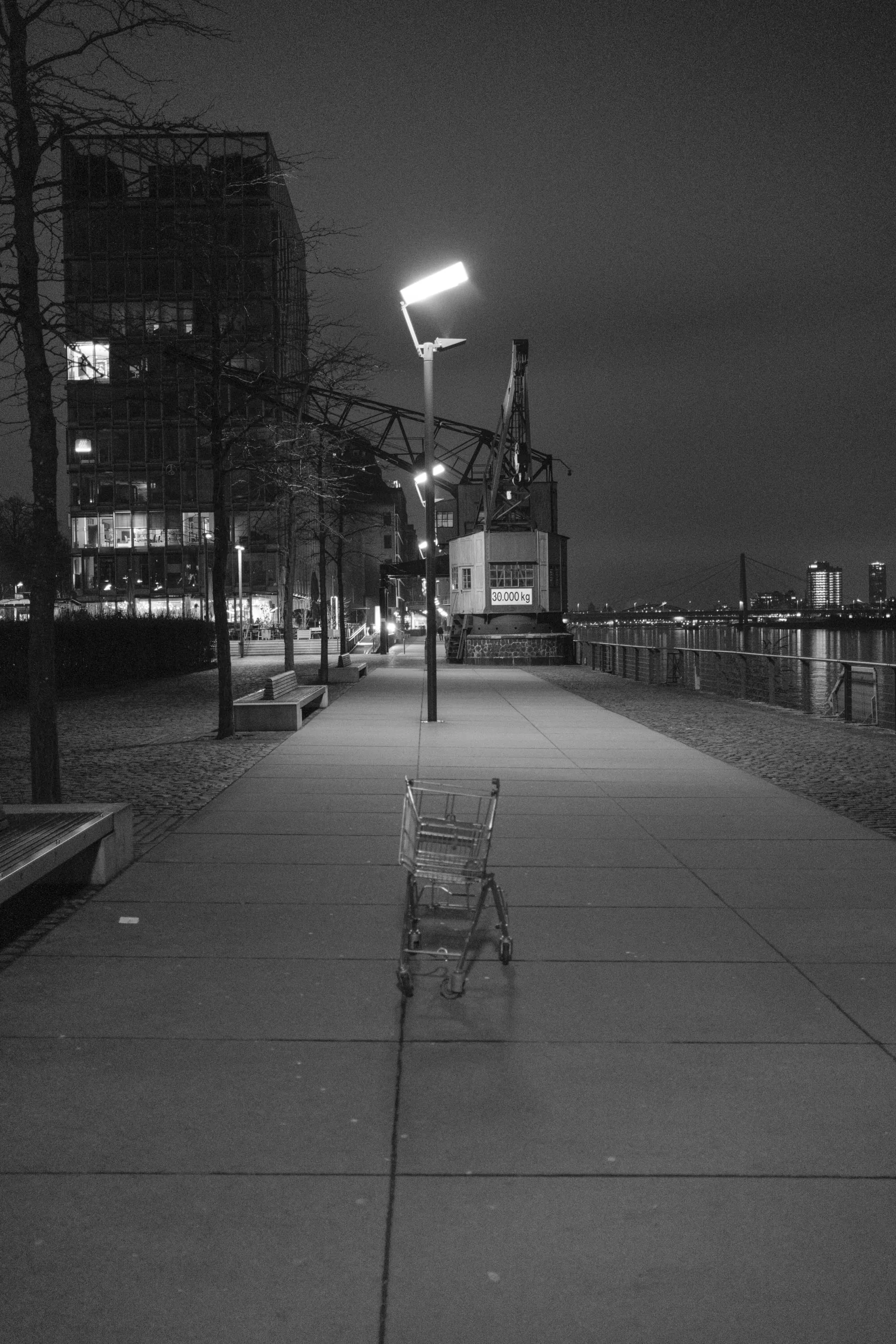 Empty shopping cart on a dimly lit sidewalk at night, surrounded by urban landscape and lampposts.