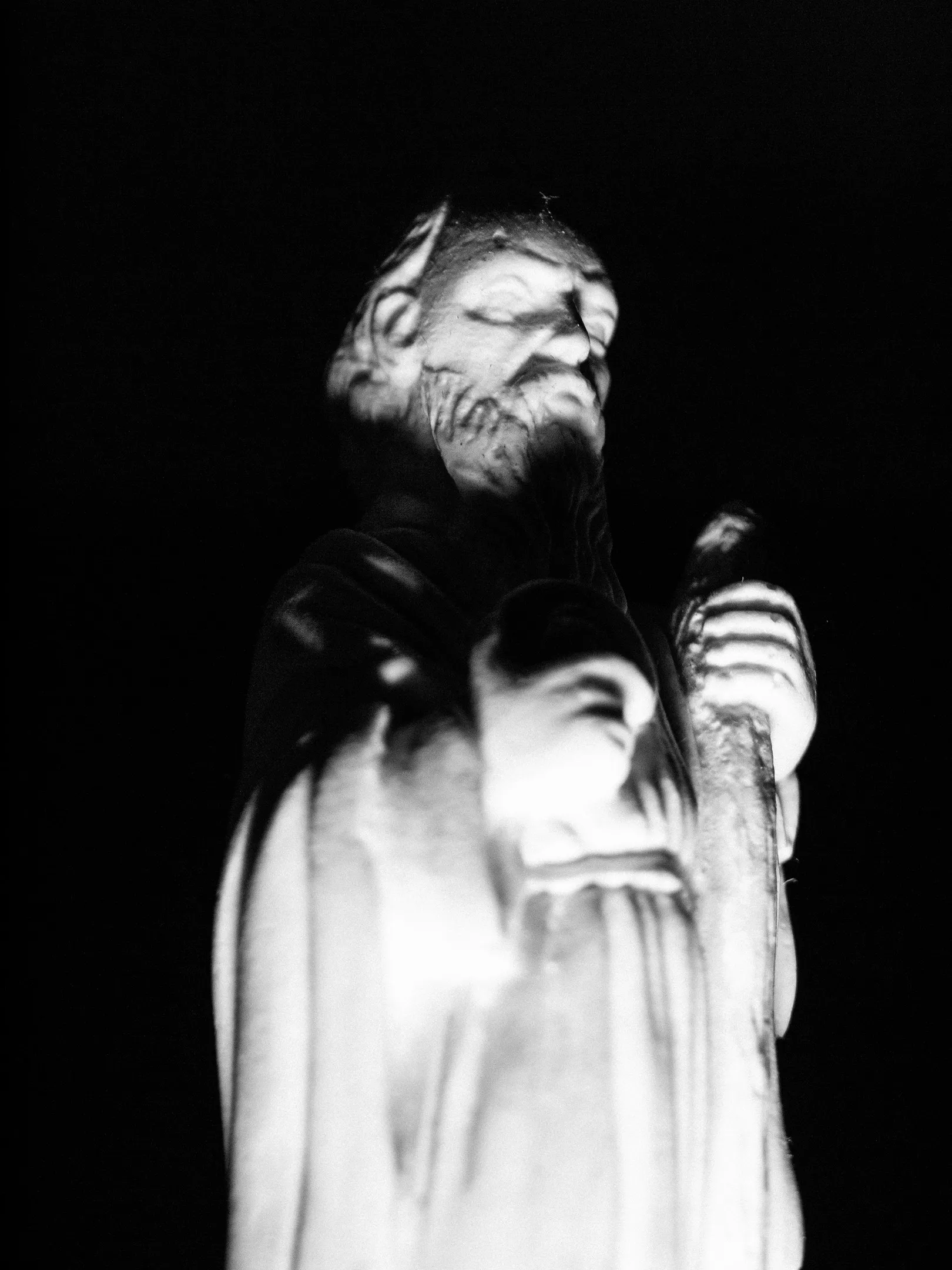 A man with long hair and beard, holding a microphone on stage, illuminated by spotlight against dark background.