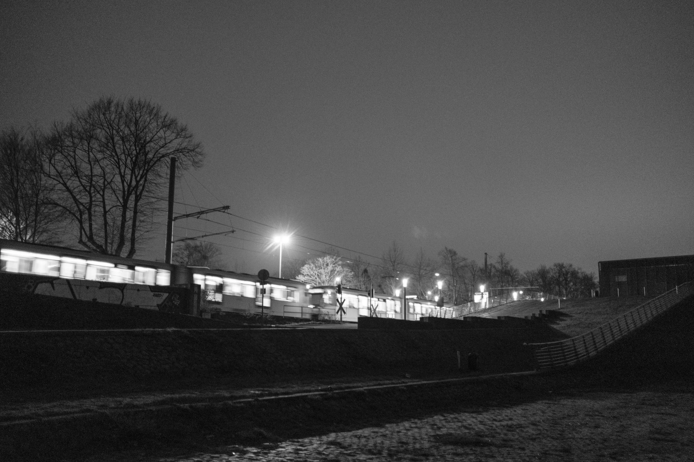 Nighttime scene with illuminated train on tracks, surrounded by bare trees, and a lamppost glowing in the distance.