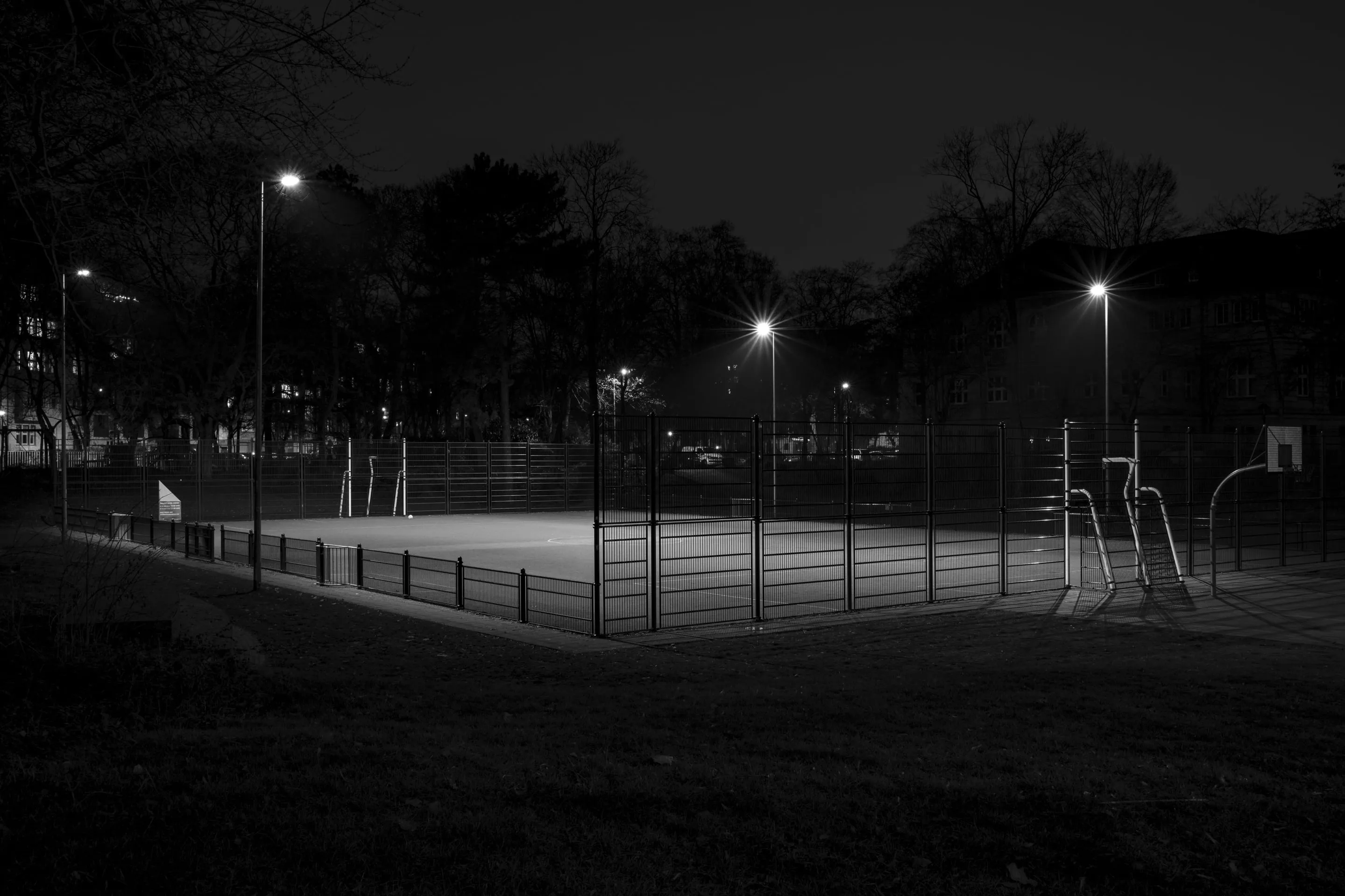 Tennis court at night, illuminated by two lampposts on a foggy evening, surrounded by trees and buildings in the background.