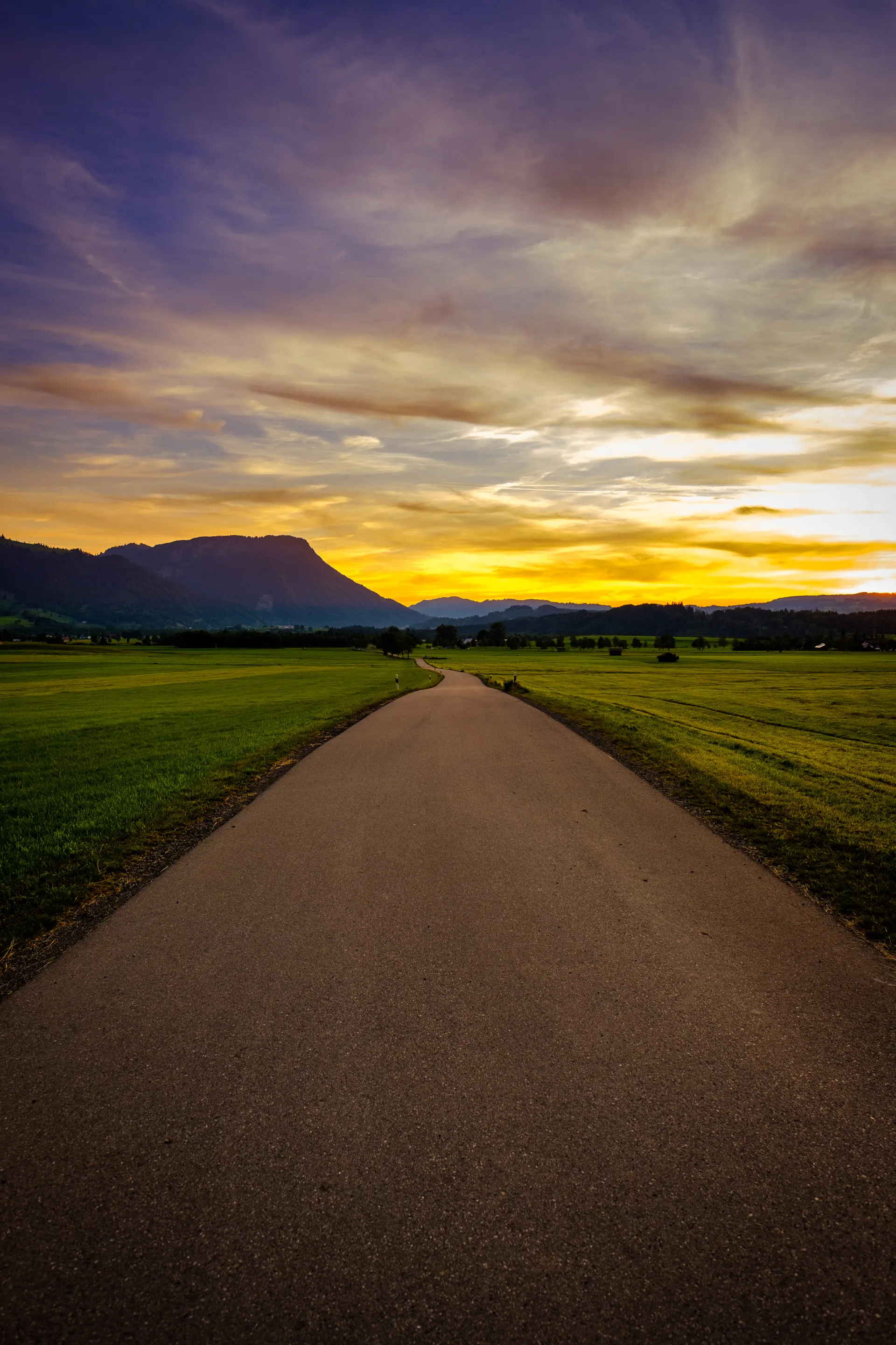 A winding road leading towards a vibrant sunset over fields and mountains.