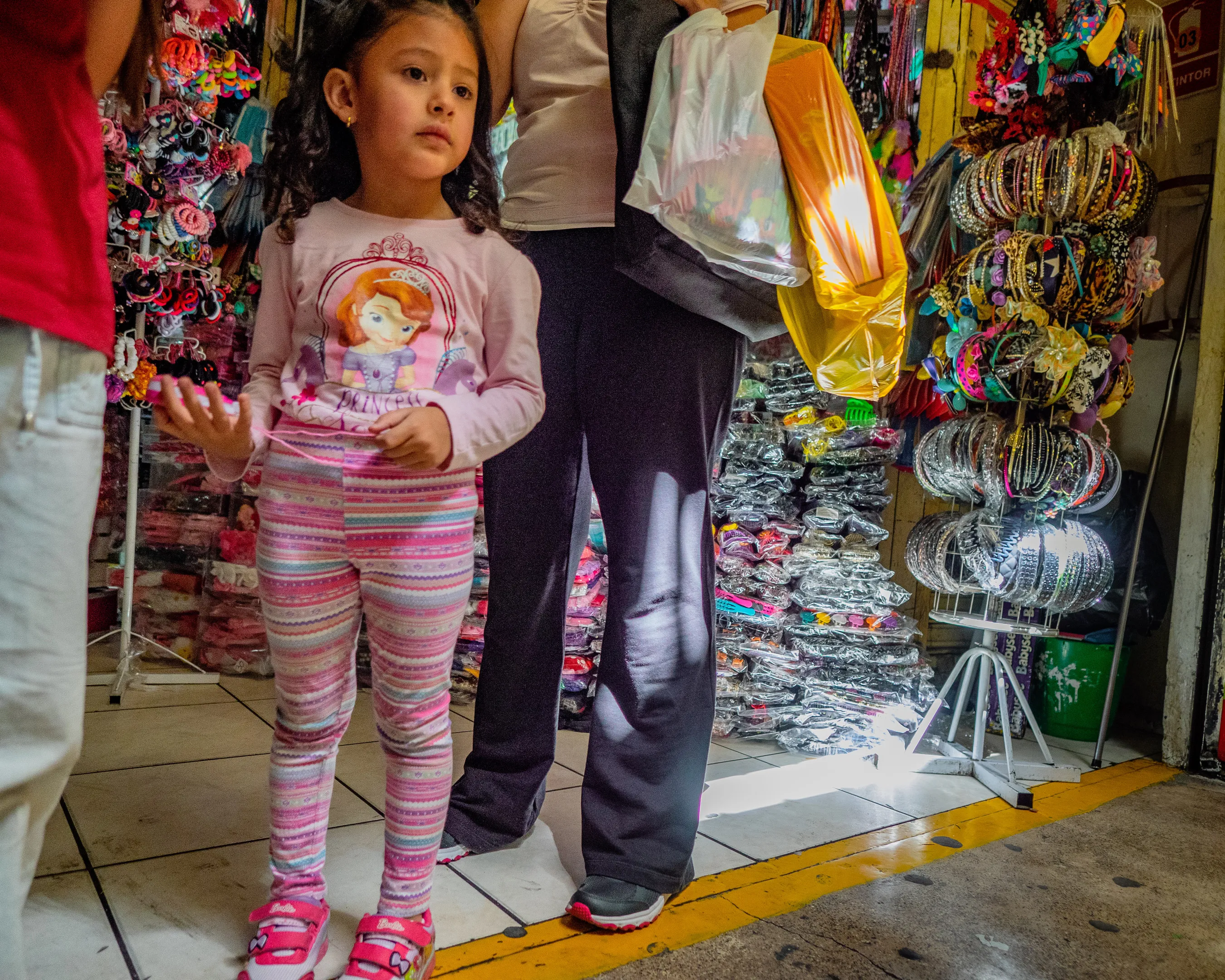 Little girl in pink striped pants and long-sleeve shirt, standing in a market with an adult.