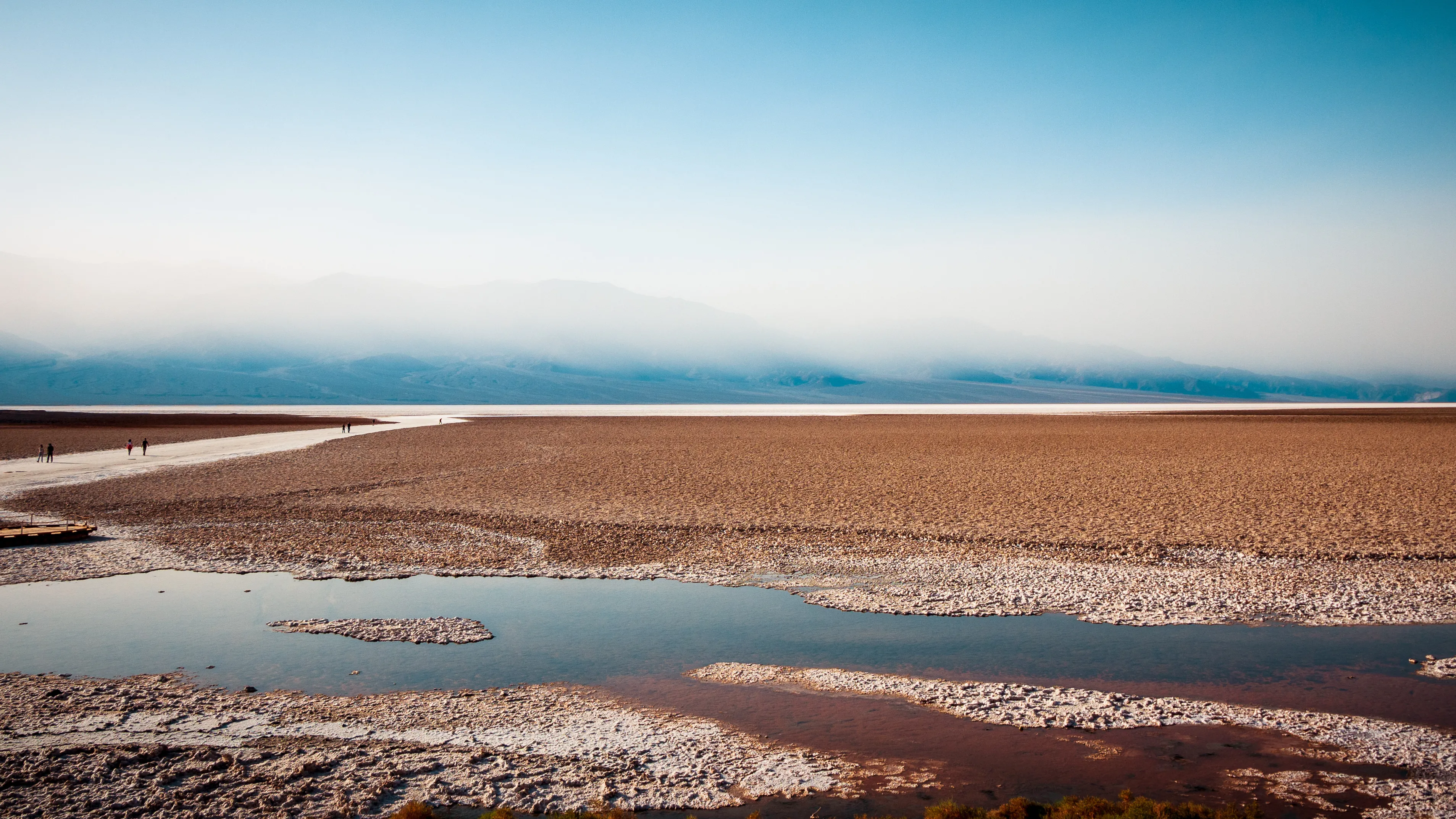A wide, shallow body of water with a pebbled shore, leading to distant mountains under an overcast sky.