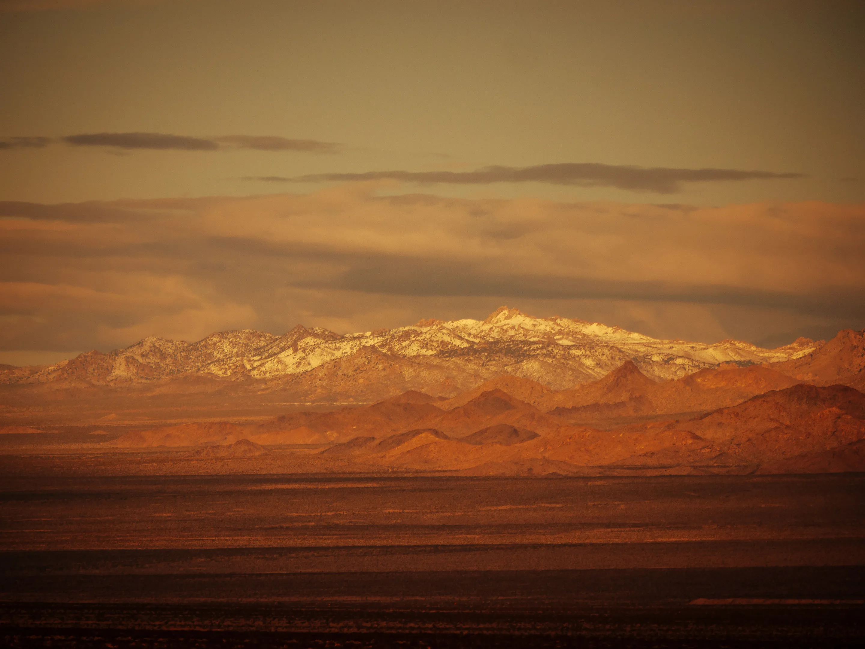 Mountain range at sunset, snow-capped peaks, warm orange hues in sky and landscape.