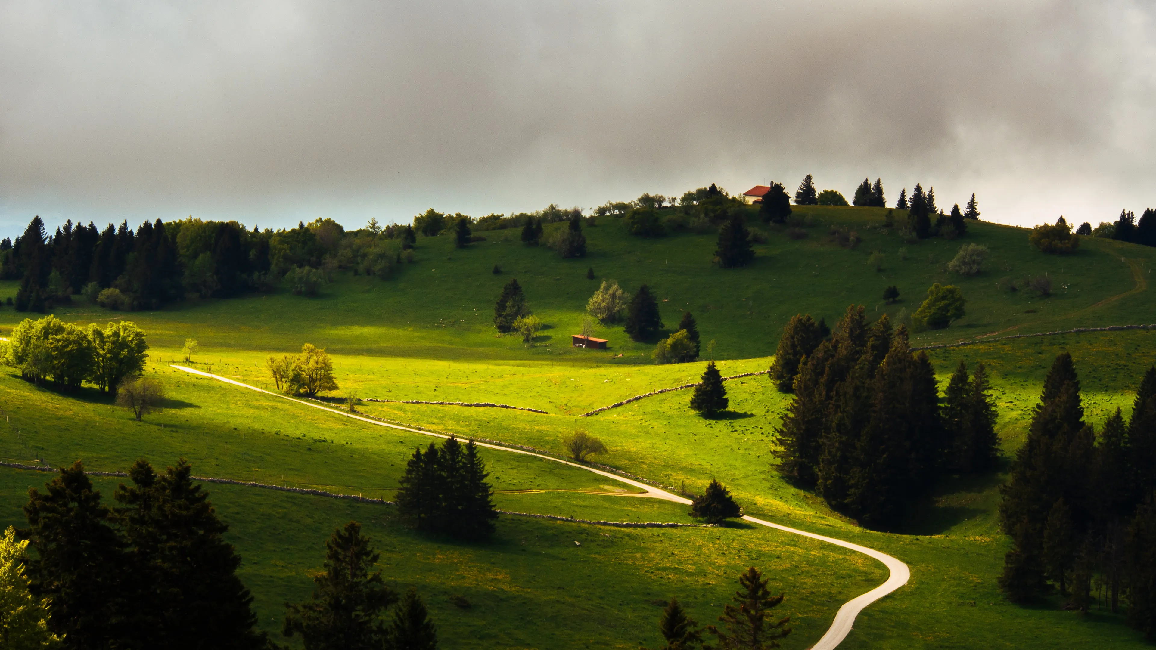 Green hills with winding path, trees, house, and cloudy sky.