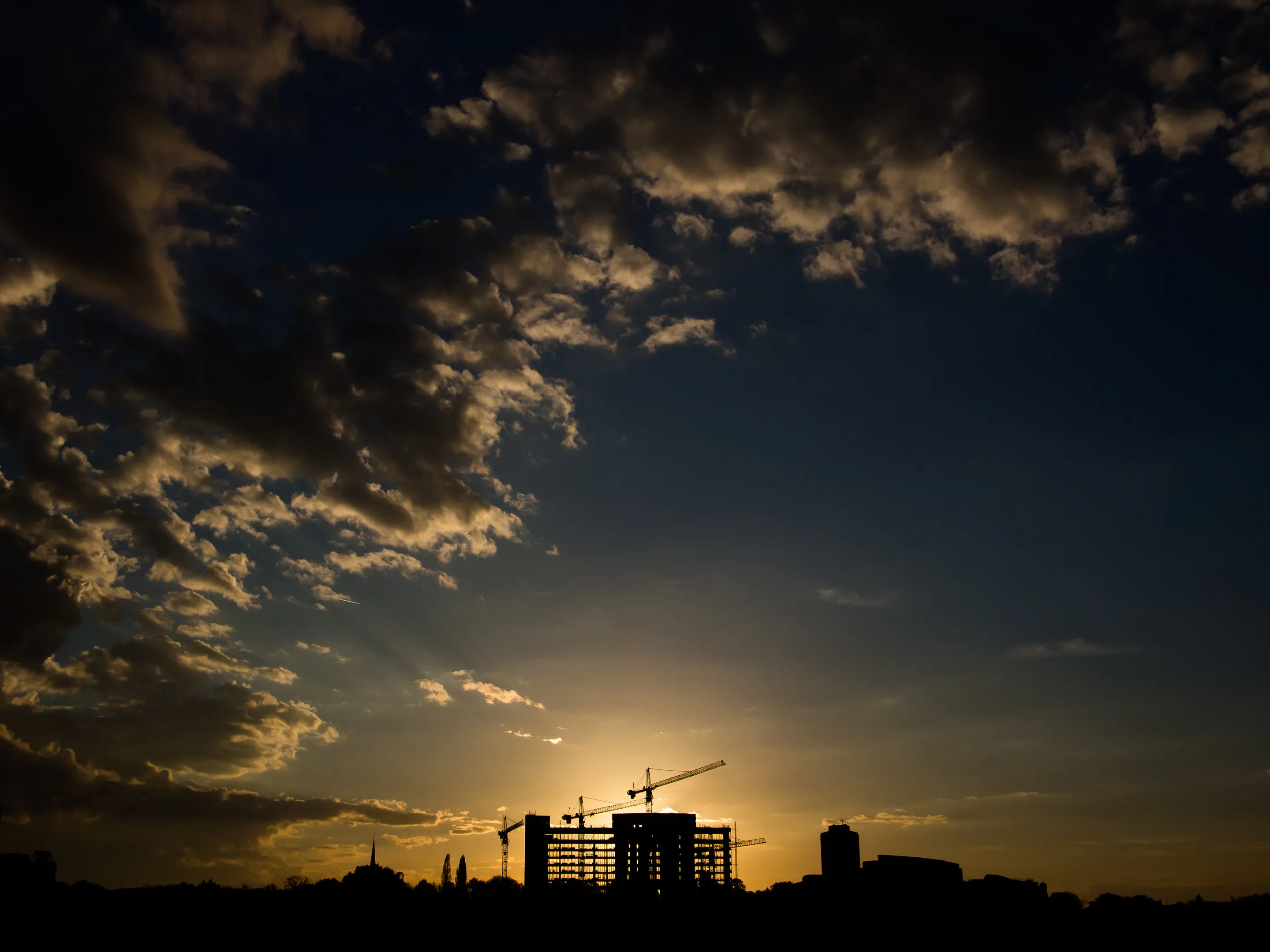 Silhouettes of cranes and a partially constructed building against a dramatic sunset sky with scattered clouds, casting shadows over the city skyline.