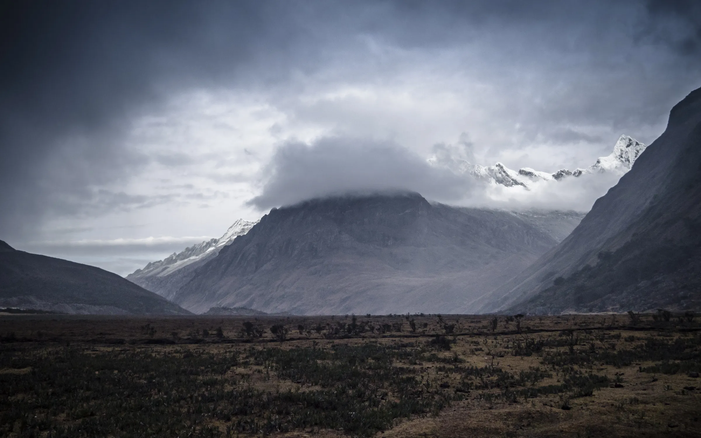 Mountain range with snow-capped peaks, partly obscured by dramatic clouds, set against a vast, barren landscape.