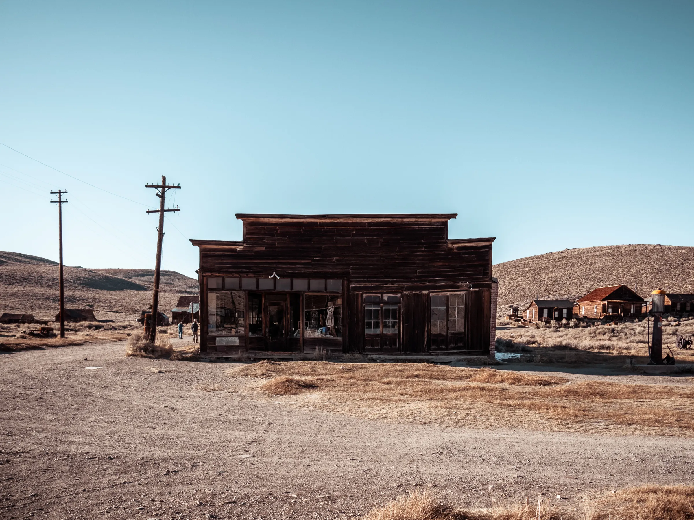 Abandoned wooden building with two front doors, surrounded by dry landscape and power lines.