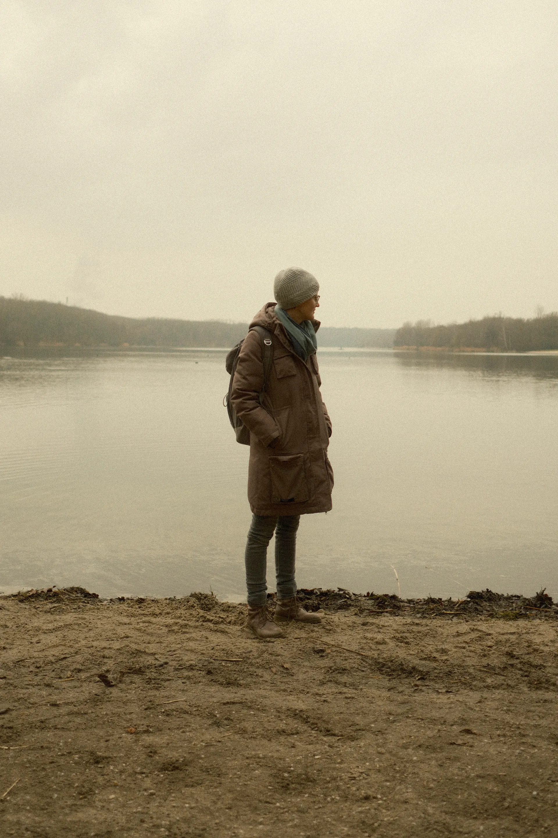 Person in coat standing by calm lake on a cloudy day.