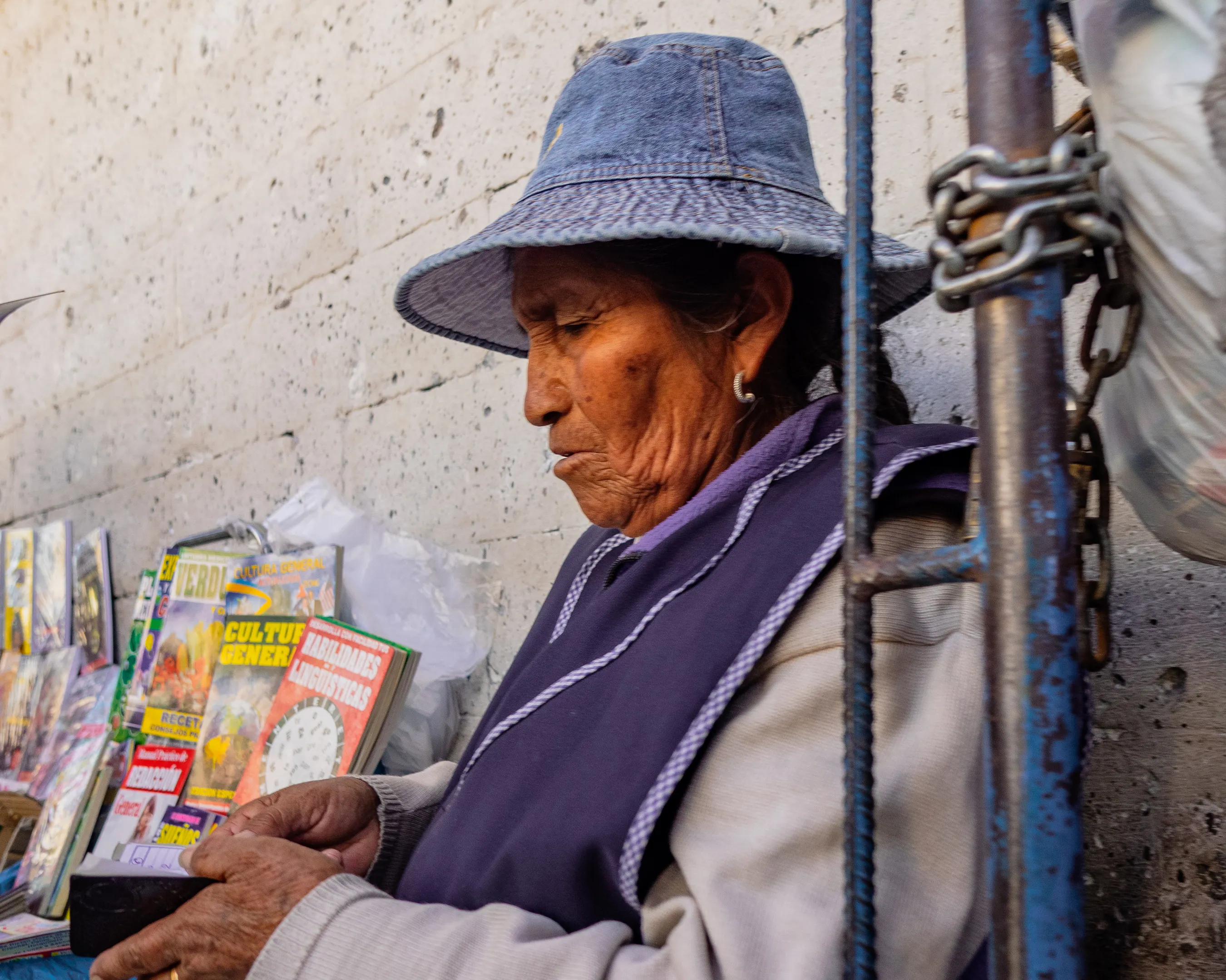 Elderly woman in blue hat and sweater, selling colorful postcards on street, next to metal gate with chain.