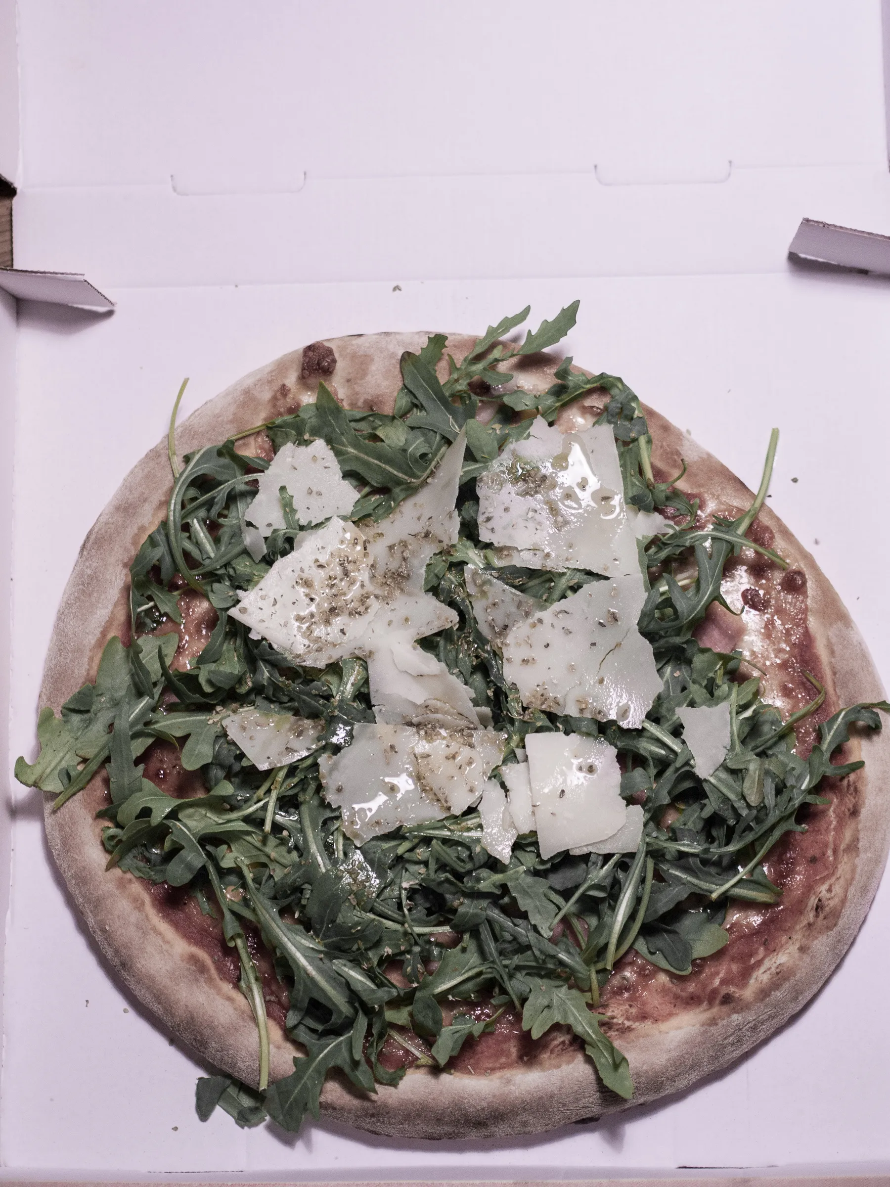 Pizza with arugula leaves and shaved cheese on a white box.