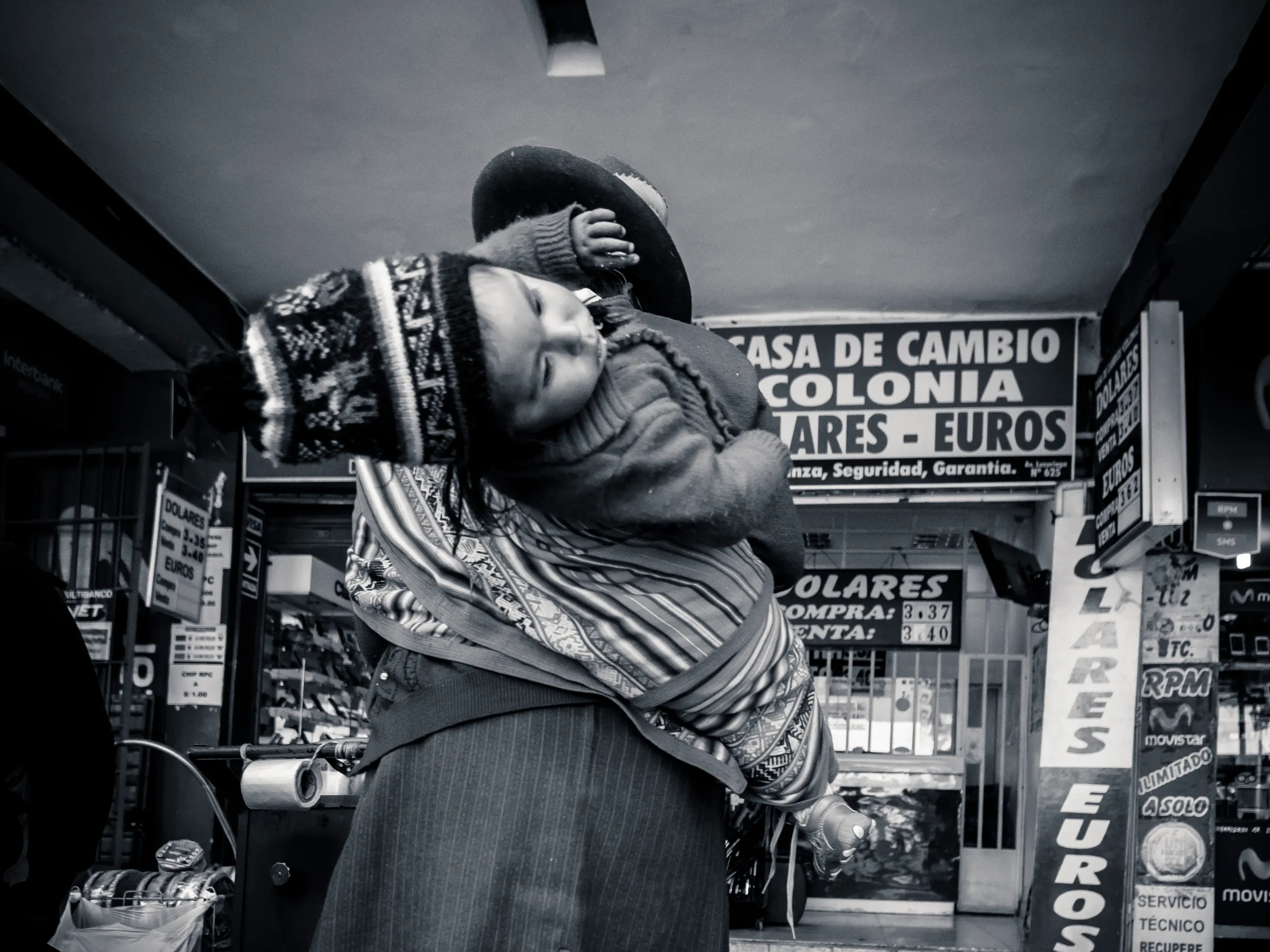 Woman carrying child on her back in front of a currency exchange store, both looking down.