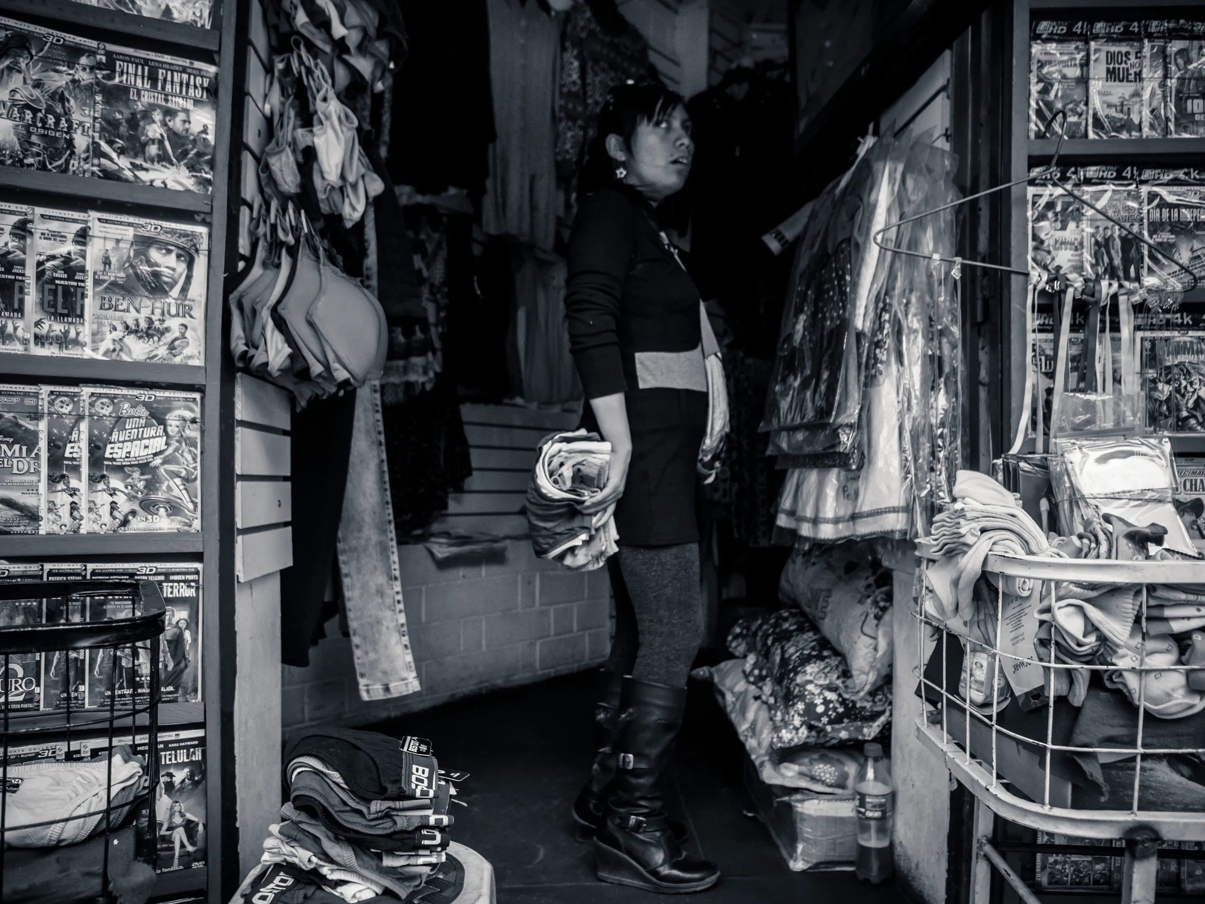 Woman in a cluttered market stall holding a bowl, surrounded by various items and framed pictures on walls.