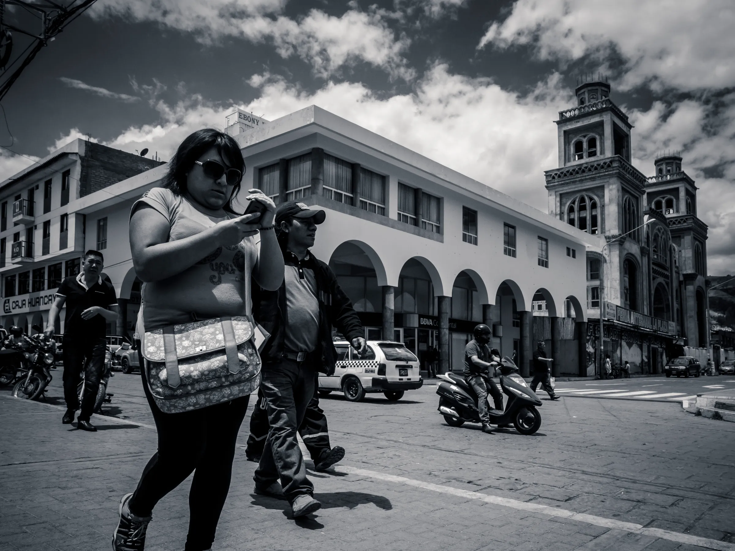 Woman adjusting sunglasses on bustling street with historic architecture in background, black and white photo.