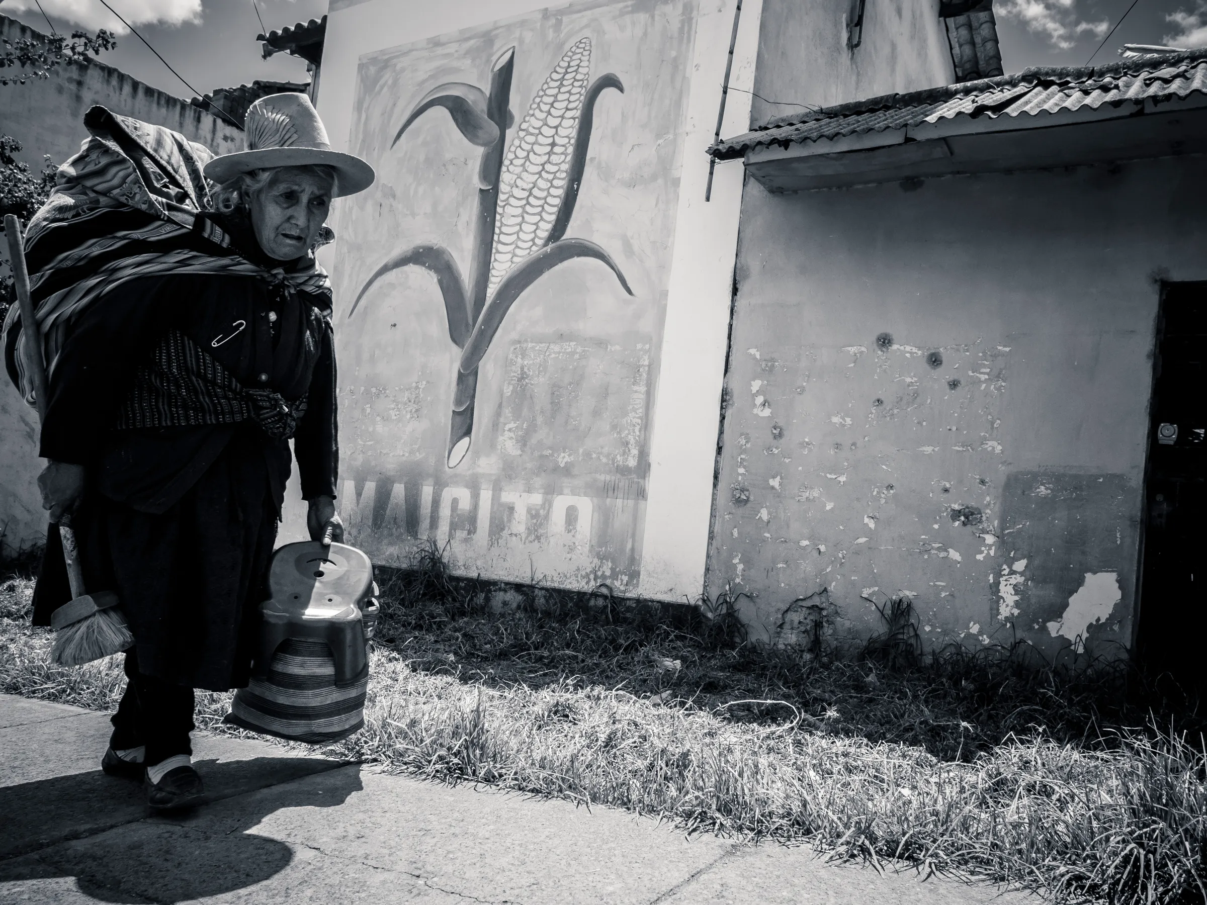 Elderly woman in traditional hat carrying a bucket on a street with a corn mural.