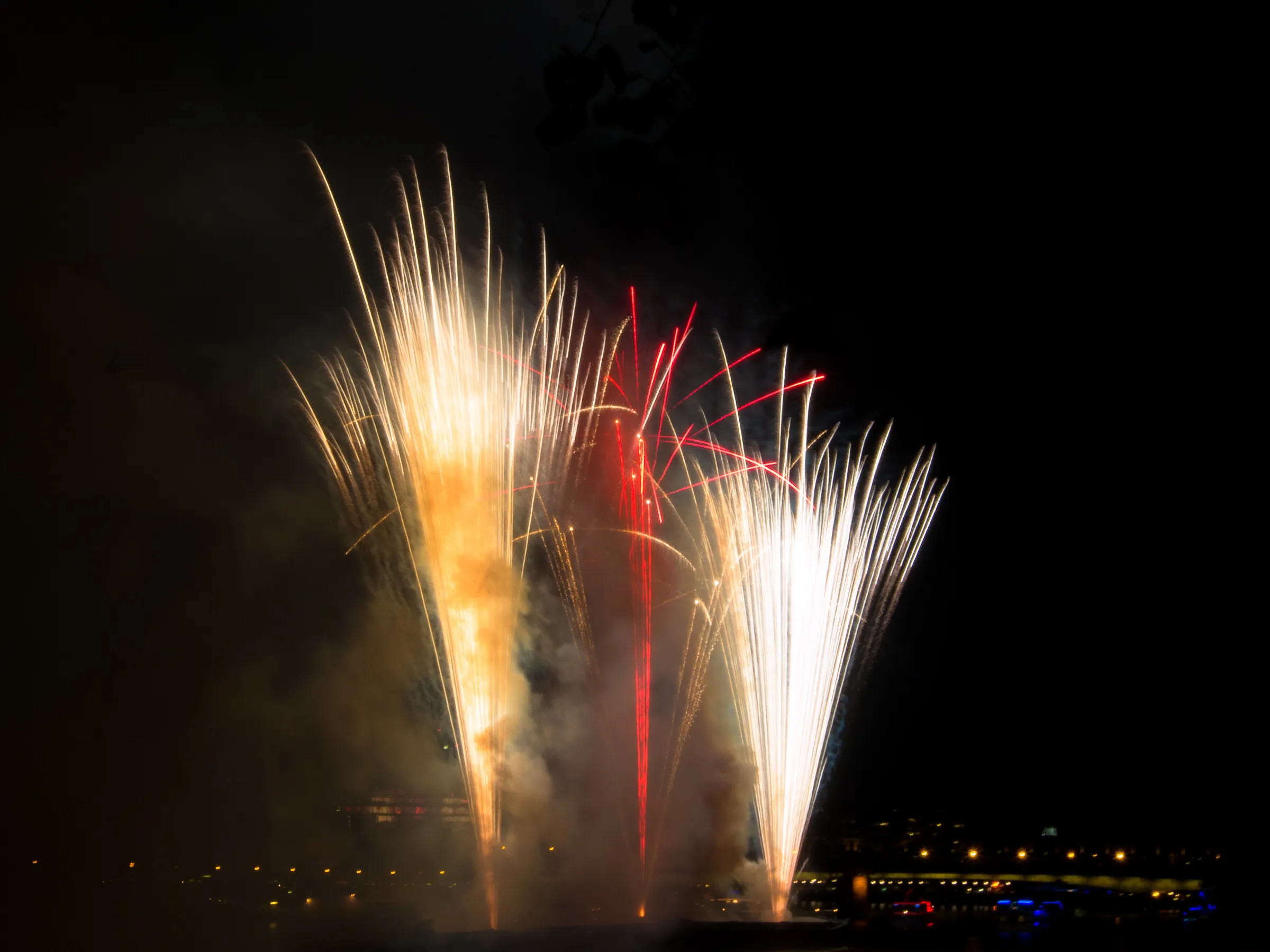 Colorful fireworks bursting in night sky. Vibrant reds, yellows, and whites. Dark silhouette of city in background.