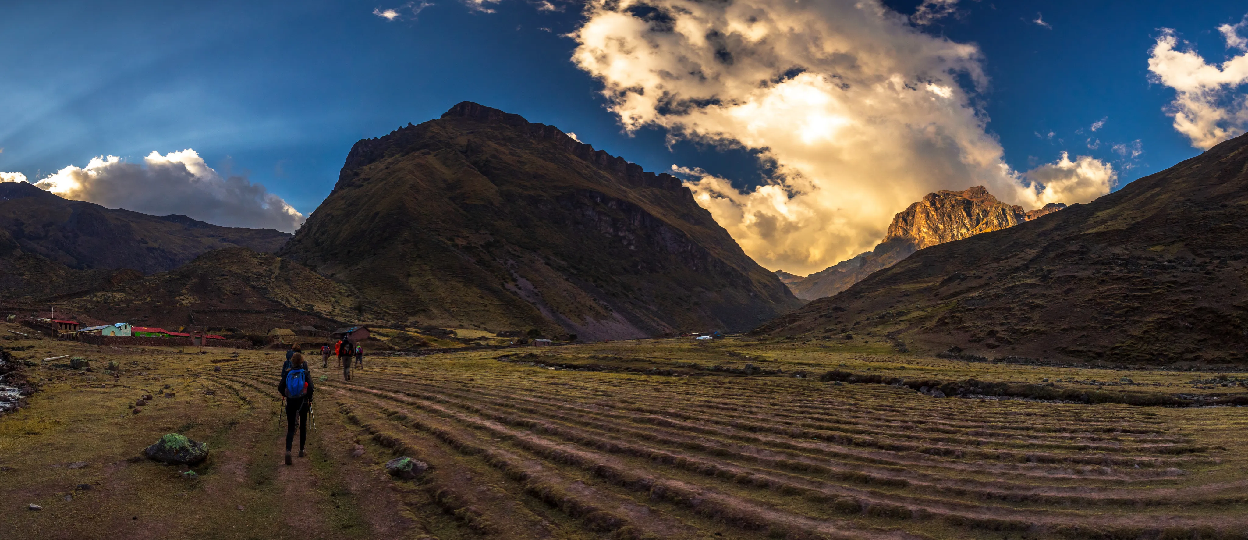 Two hikers on a dirt path in a valley, with dramatic mountains and cloudy sky in the background.