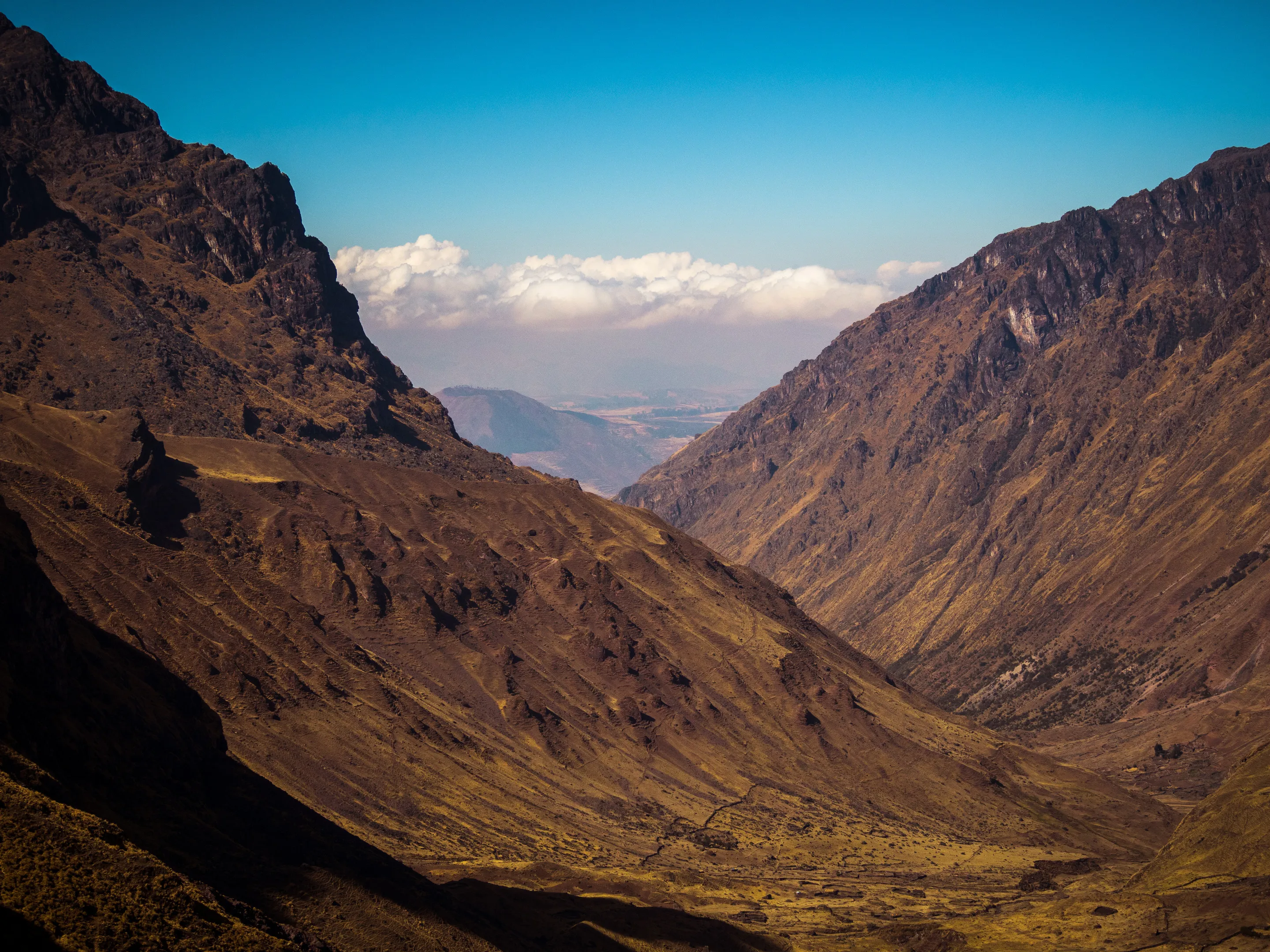 Mountain valley with rugged cliffs, golden grass, and cloud-covered peaks under blue sky.