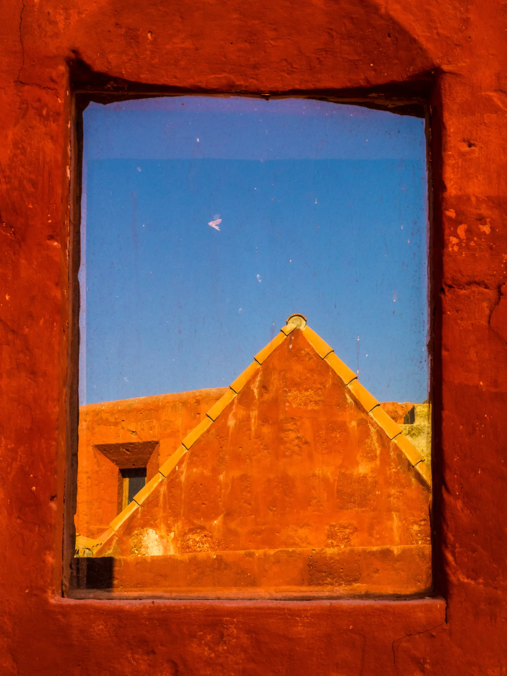 A bird flying in clear blue sky seen through a rustic window frame, with a view of earthy rooftops.