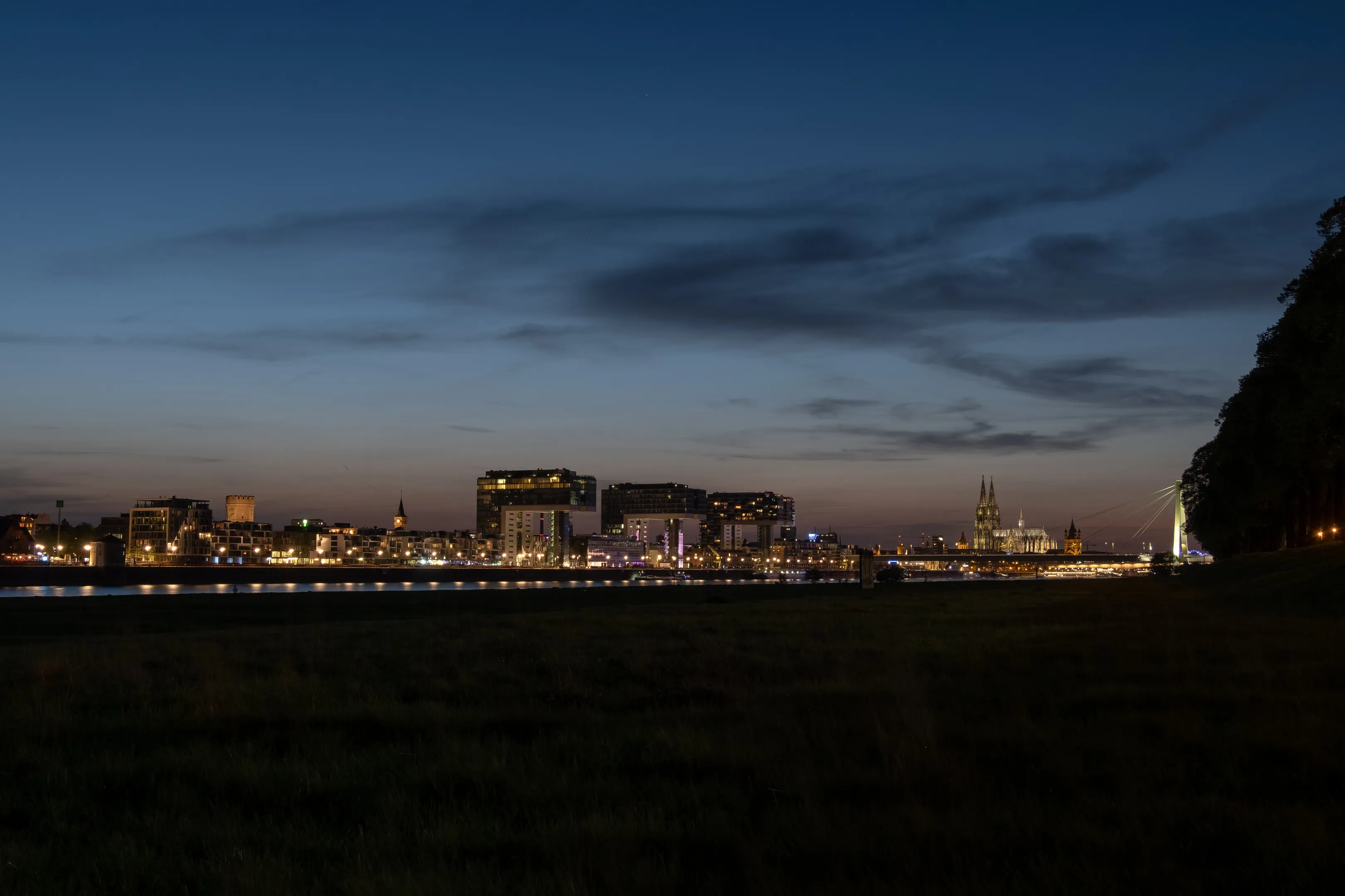 City skyline at dusk, buildings illuminated against dark blue sky with scattered clouds, water in foreground.