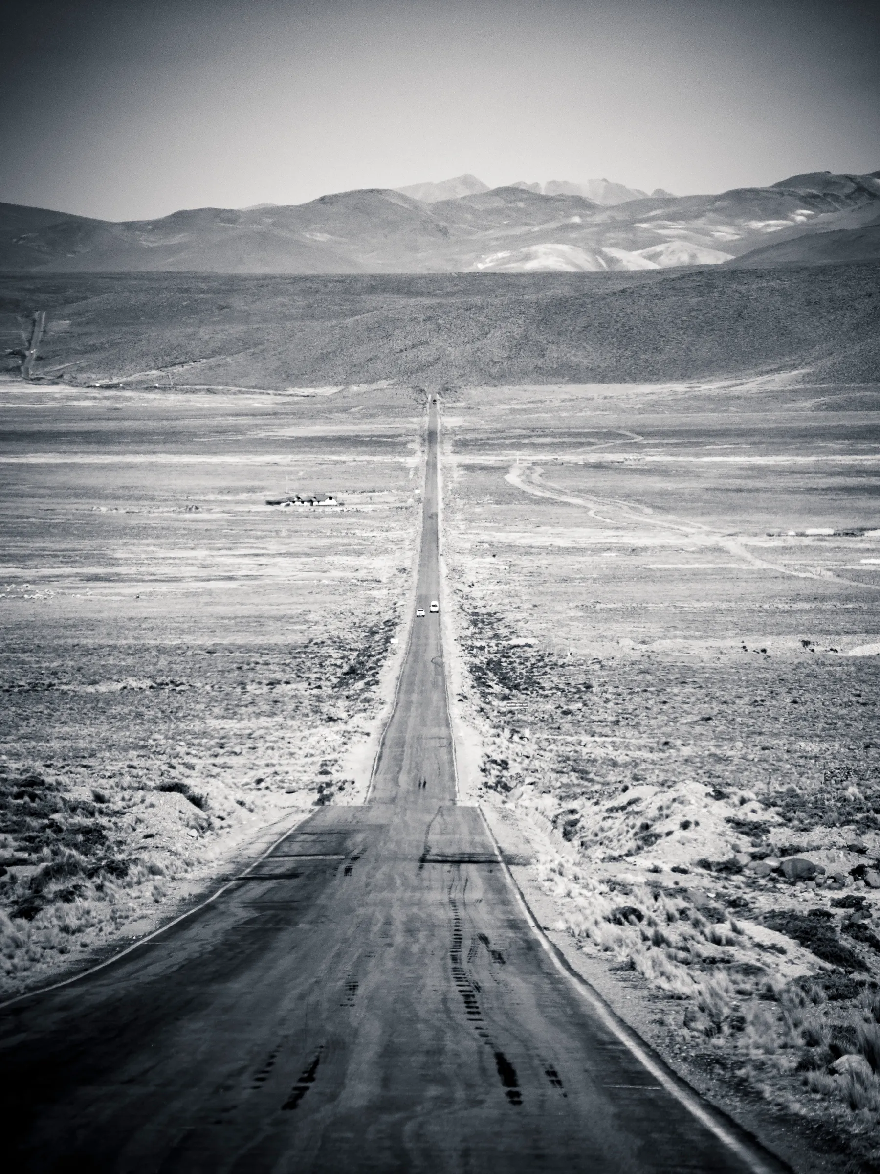 Lone car on long, straight, snowy road in desert, mountains in background.