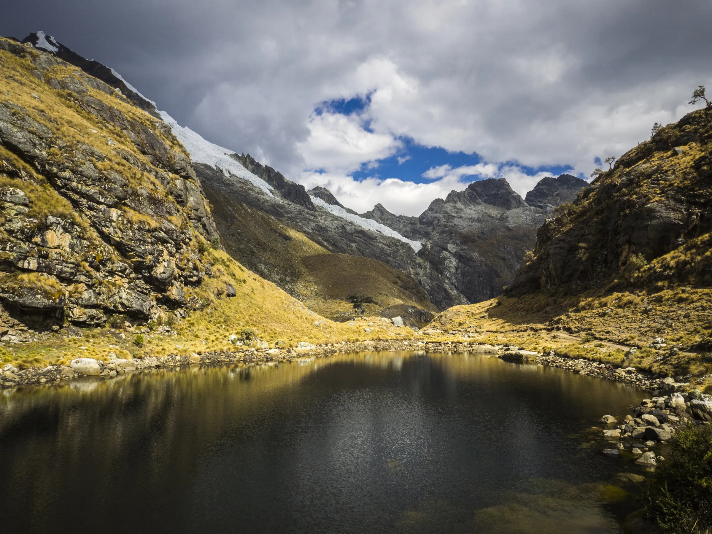 Mountain valley with snow-capped peaks, cloudy sky, and a serene lake reflecting the landscape.