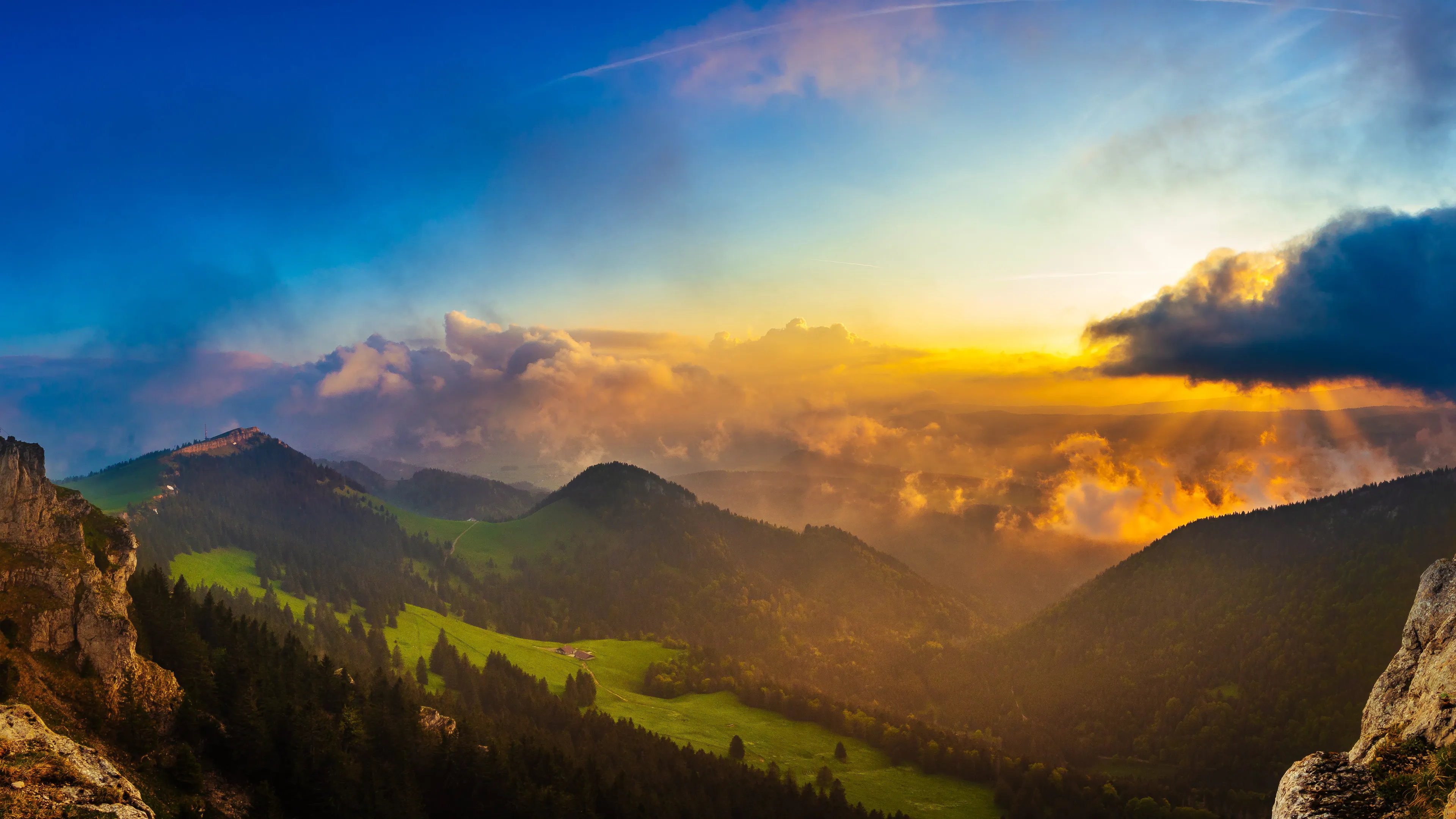 Sunset over mountains with golden light, green valleys, and dark clouds.