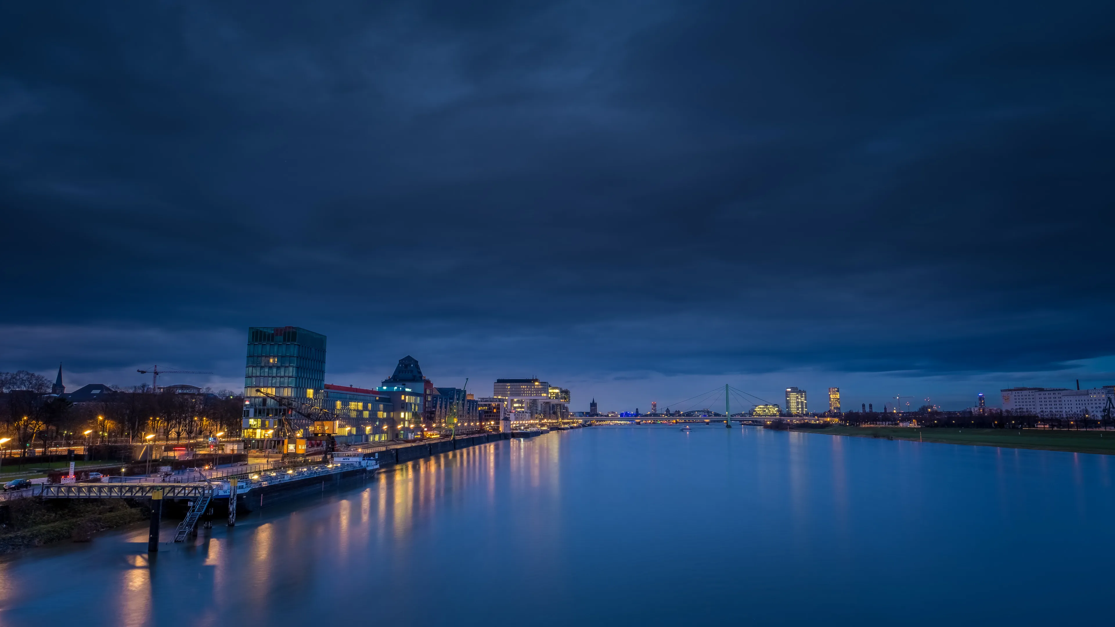 City skyline at dusk, buildings illuminated, river reflecting lights, cloudy sky.