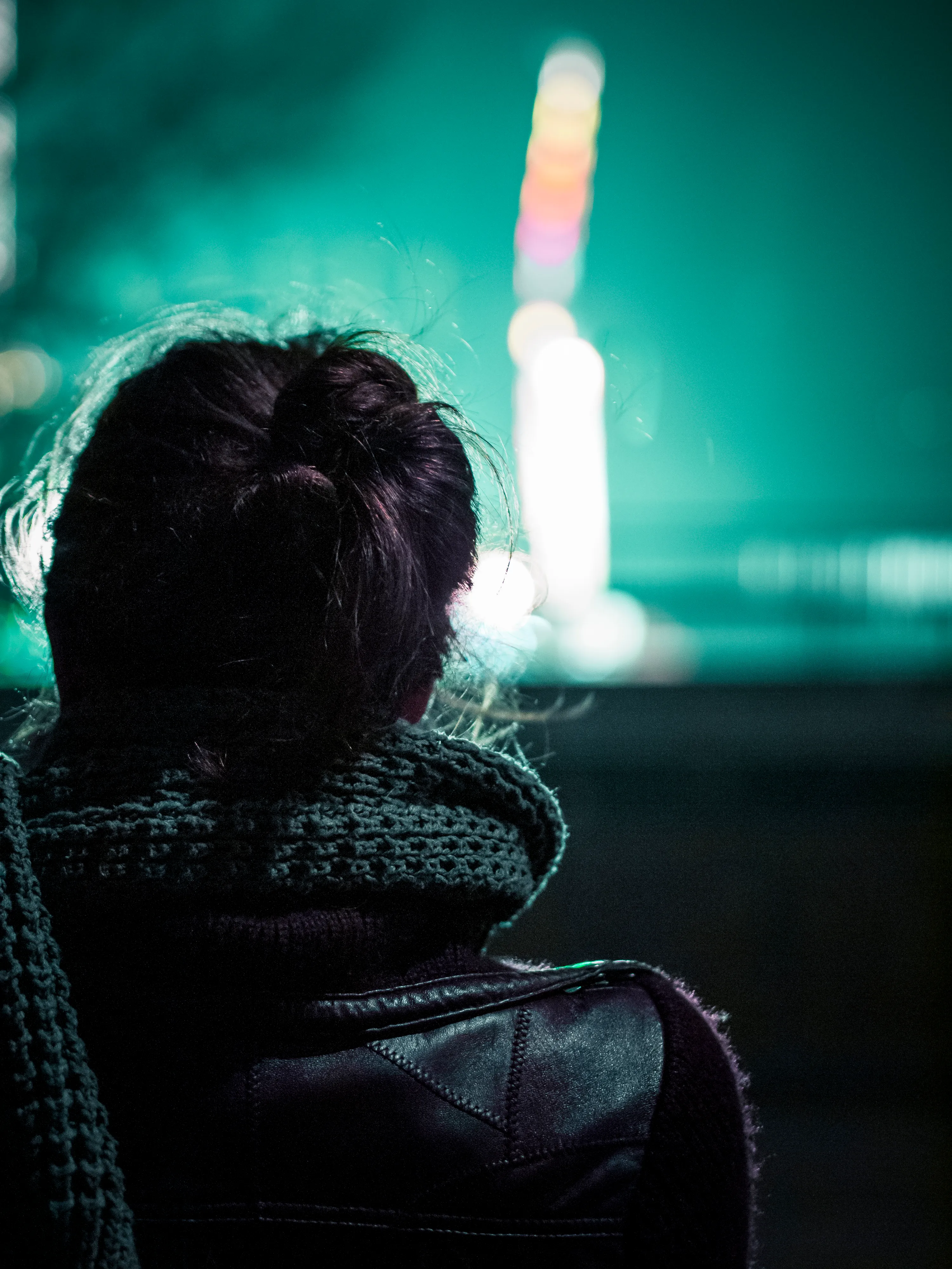 Person with dark hair in bun, wearing jacket, facing away from camera at night with blurred lights background.