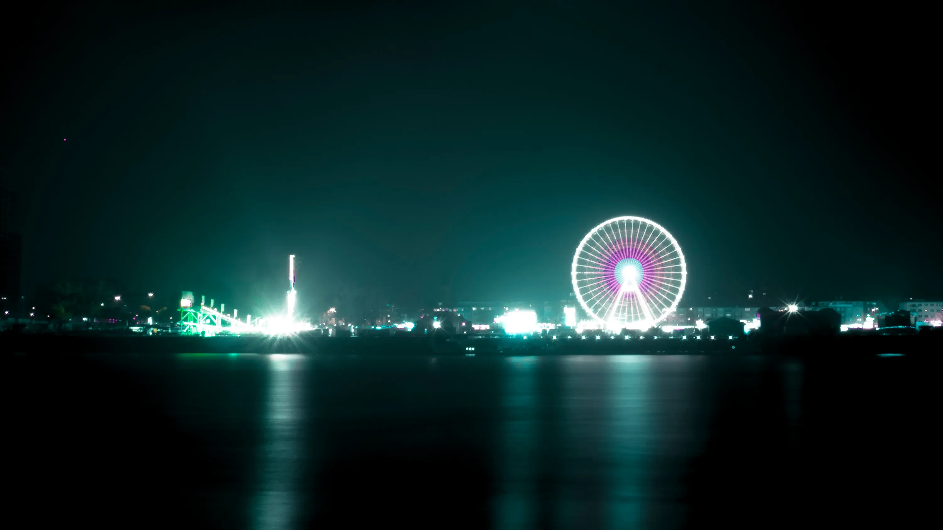 Night view of illuminated Ferris wheel and city skyline reflected on calm water, with a lit tower in the distance.