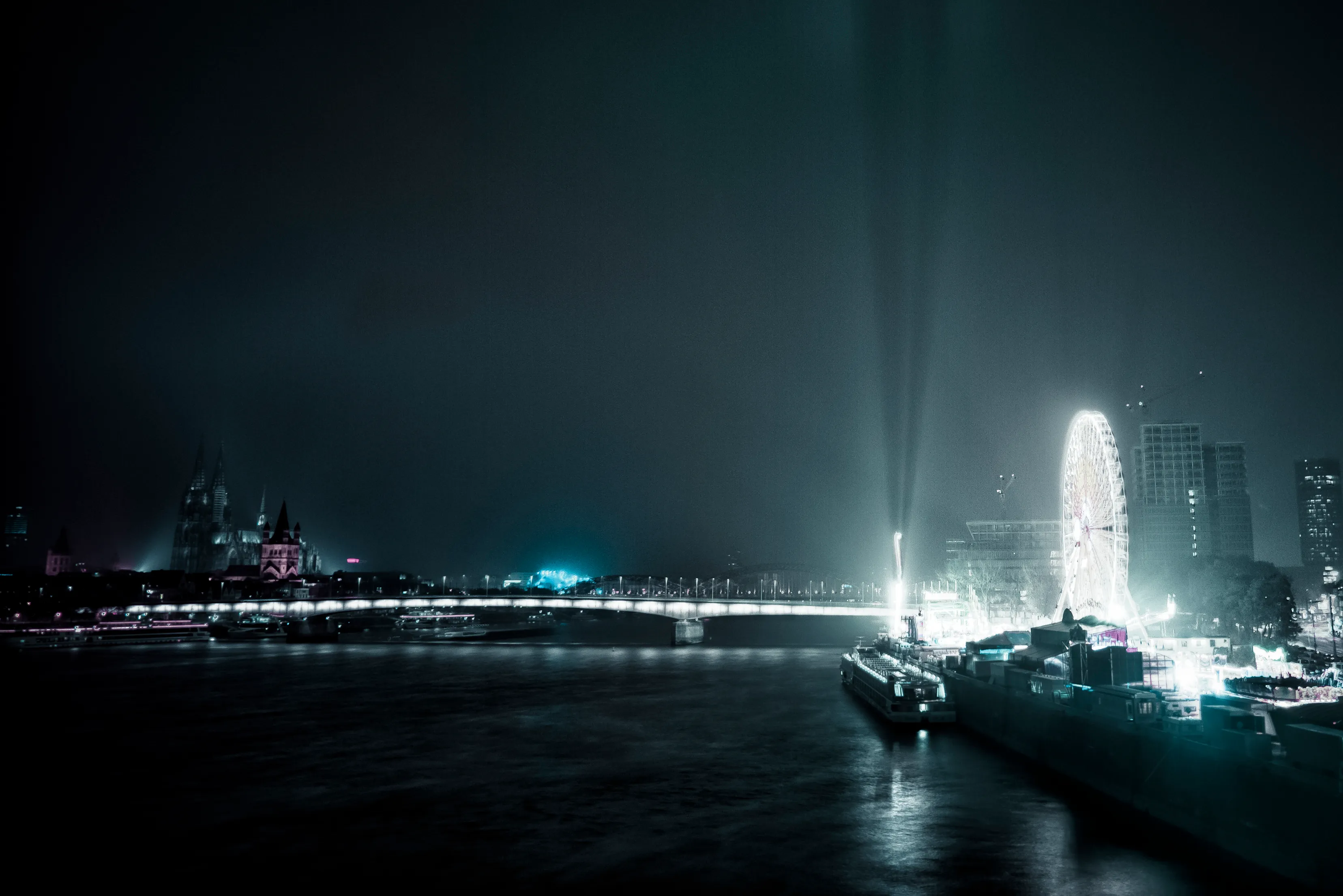 Nighttime cityscape with illuminated Ferris wheel, buildings, and searchlight beams over a river.