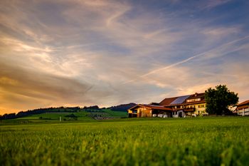Golden light on farmhouse in lush green field with dramatic sunset sky and distant hills.