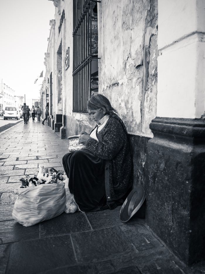 Woman sitting against wall on street, holding bread, with a bag of items beside her.