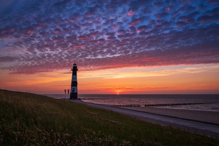 Lighthouse at sunset with vibrant clouds over a calm sea, grassy foreground.